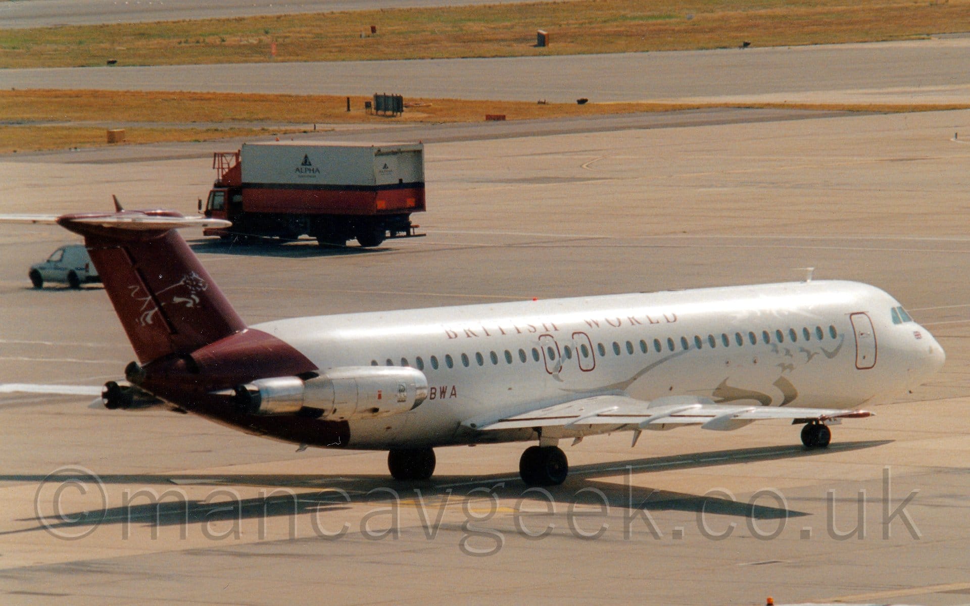 Side view of a twin engined jet airliner with the engines mounted on the sides of the rear fuselage, taxiing from left to right. The plane is mostly white, with the outline of a silver lion on the forward fuselage, and maroon "British World" titles on the upper fuselage. The rear fuselage and tail are maroon, with a smaller silver lion on the tail. Grey concrete apron and taxiways fill most of the rest of the frame, with strips of grass separating taxiways in the background, as well as a pink and white truck driving from right to left on the left of the frame.