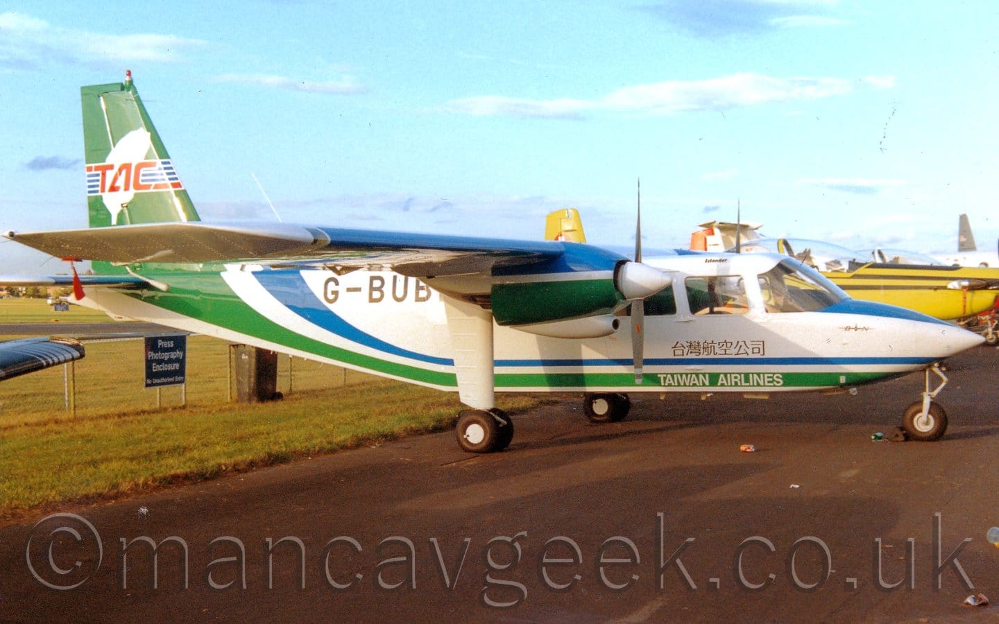Side view of a very small, high-winged, twin propellor-engined airliner parked on black tarmac facing to the right. The plane is mostly white, with a blue and green stripe running along the belly, sweeping up in to the tail. There are small white "Taiwan Airlines" titles on the green stripe under the cockpit, with some black writing in a Chinese script above it. The registration "G-BUB_" is on the upper rear fuselage in black, the last letter obscured by the fixed landing gear coming down from the green and blue engine pods on the wings. A black tarmac taxiway fills most of the foreground, lined with grass on the left of the frame. The black tip of a planes wing is peeking in to frame on the left, while the nose and cockpit of a yellow 2-seat trainer is in the background on the right. Pale blue sky with fluffs of white cloud fill the rest of the frame.