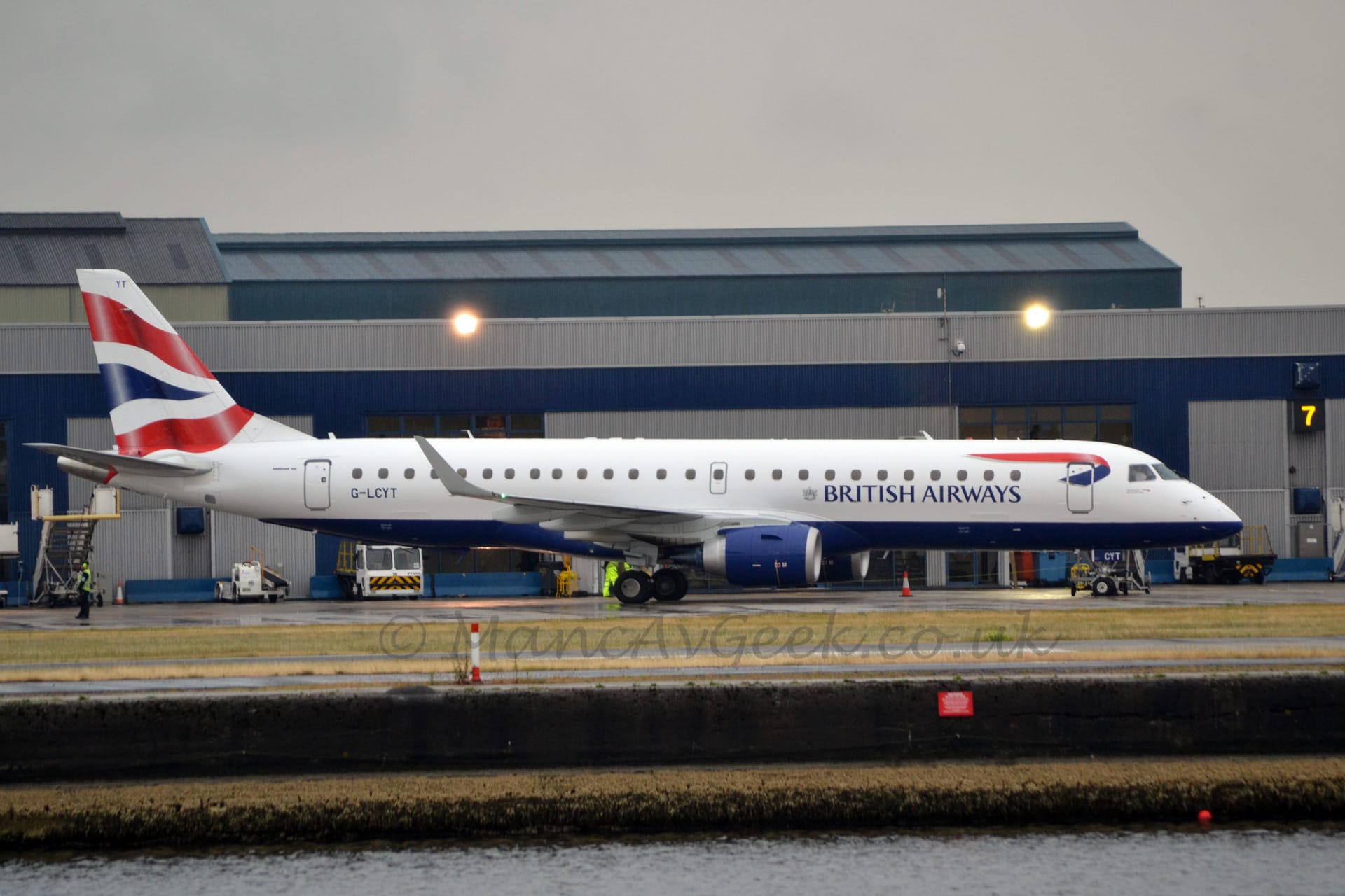 Side view of a twin engined jet airliner taxiing from left to right. The plane is mostly white, with a dark blue belly, and dark blue "British Airways" titles on the lower forward fuselage. There is a twisted, red and blue ribbon on the upper forward fuselage, directly aft of the cockpit windows, and a wavy blue stripe between a pair of wavy red stripes on the tail. The black registration "G-LCYT" is on the lower rear fuselage, under the last of the rear cabin windows. A large body of water site in the foreground, with the airport built a couple of metres above it. A grey and black terminal building stretches across the background, under grim grey skies.