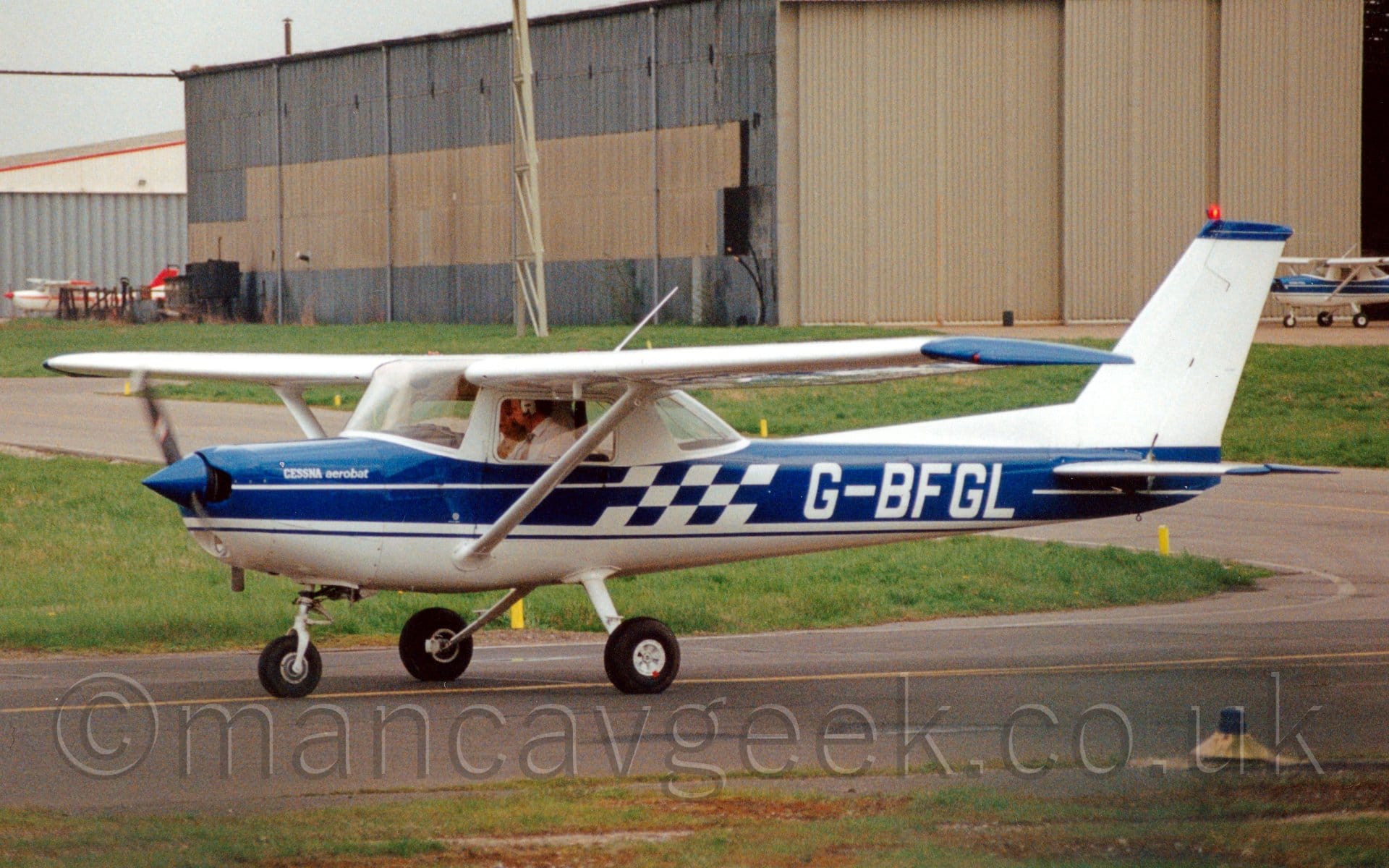 Side view of a high-winged, single-engined light aircraft taxiing from right to left. The plane is mostly blue, with a white tail and belly, as well as the frames around the cockpit windows being white There is a blue and white checker-board pattern n the middle of the fuselage, just ahead of the white registration "G-BFGL". A large grey hangar dominates the background, on the right of the frame, with grey sky visible beyond that.