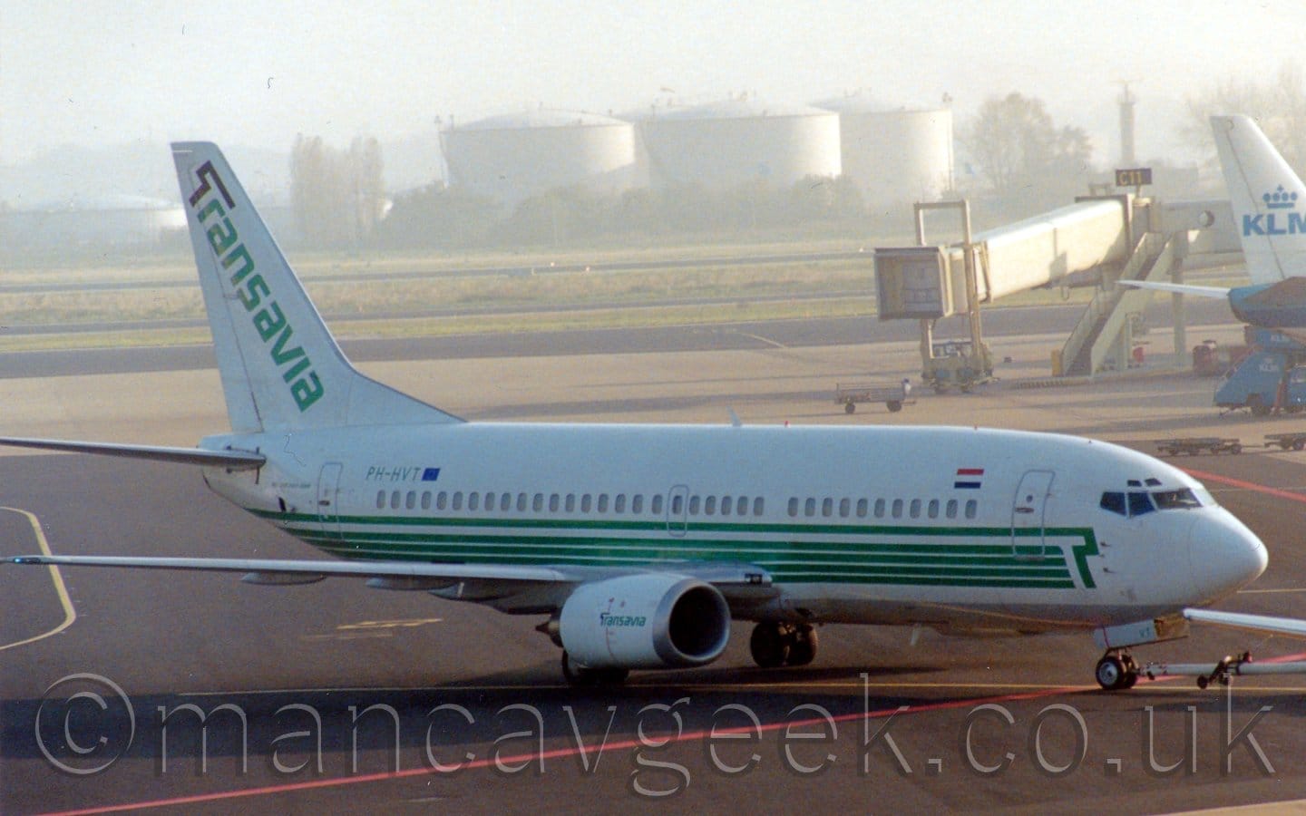 Side view of a twin engined jet airliner facing to the right but being pushed back to the left by an out-of-frame tug attached to the nose-wheel by a long white pole. The plane is mostly white, with a series of green stripes running along the lower body, negative space on the forward fuselage forming an upper-case letter "T". There are large, green, diagonal "Transavia" titles on the tail, the negative space of the "T" partially outlined in black. There are similar but smaller titles on the sides of the engine pods, mounted under the wings. Black apron with red and yellow markings fills the foreground, with black taxiways and runways separated by large grassed areas in the background, with gigantic white fuel tanks in the distance, slowly vanishing in to bright hazy skies.