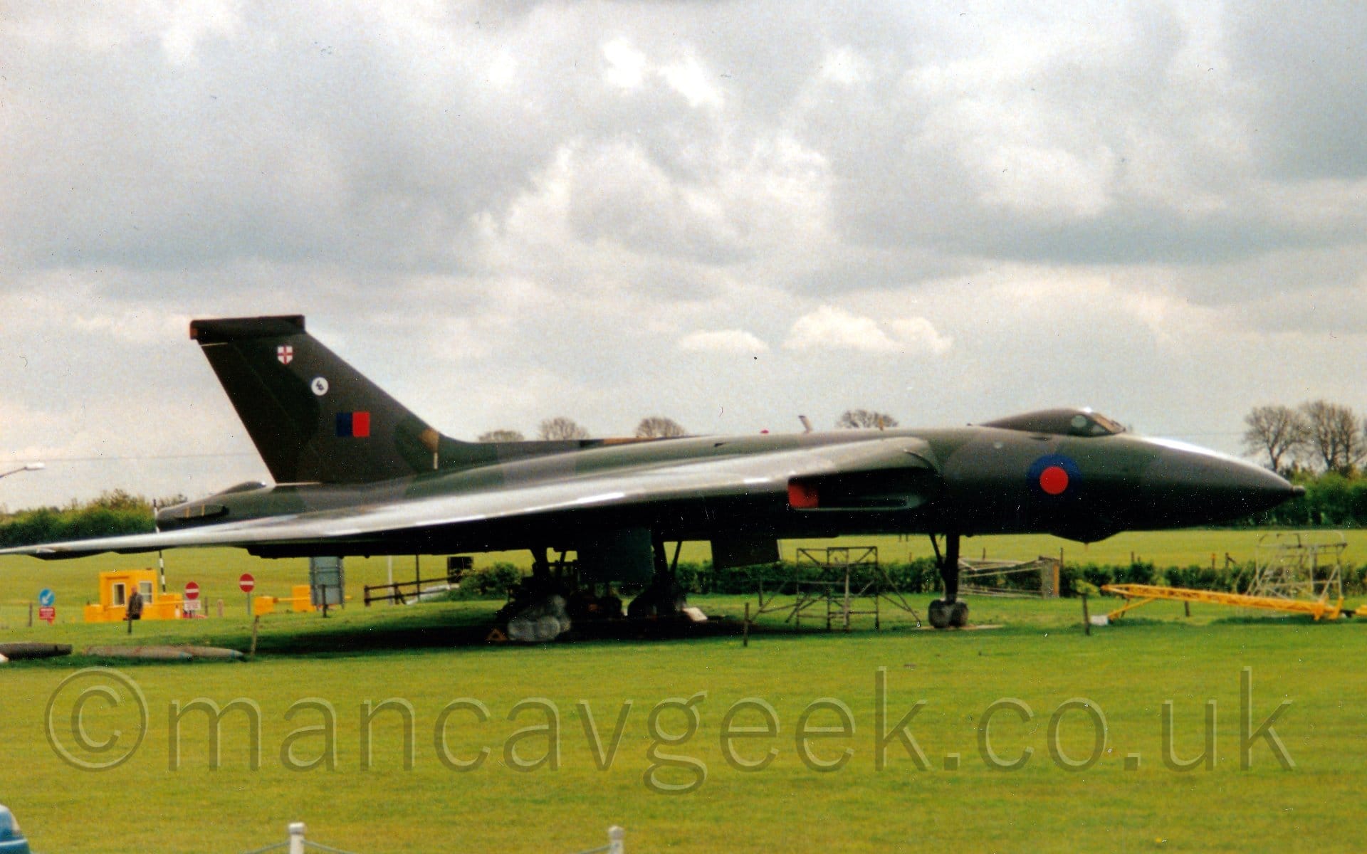 Side view of a 4 engined military jet bomber aircraft parked on concrete pads surrounded by grass, facing to the right. The plane is covered in an olive green and grey camouflage scheme, with a blue and red roundel on the forward fuselage below the cockpit, and a blue and red fin flash on the tail, below a circular blue and white logo, and a white shield with a red cross in the middle. The engines, buried in the wing roots, have red covers over the inlets. Green grass fills the foreground, with dramatic cloudy grey skies filling the rest of the frame.