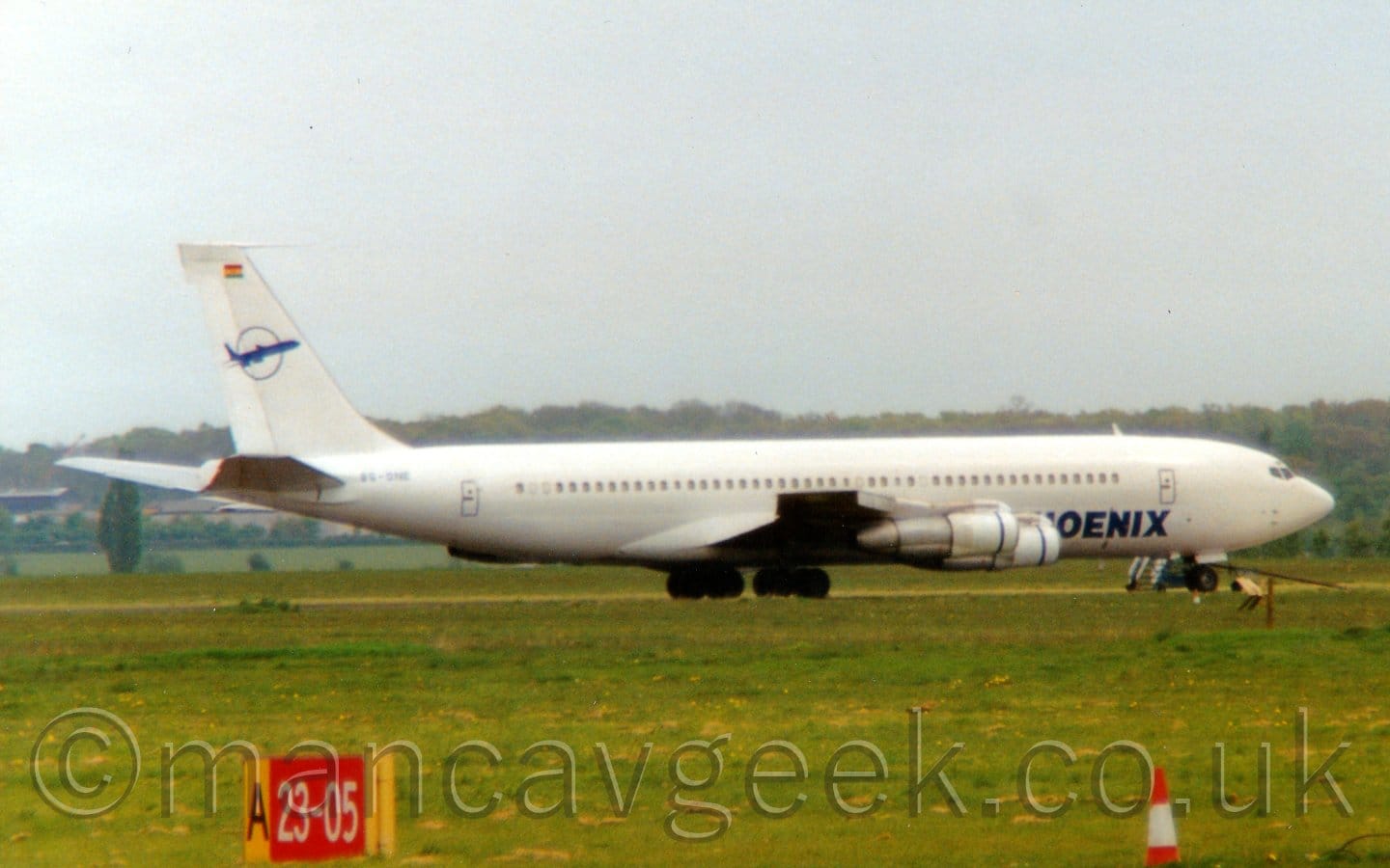 Side view of a 4 engined jet airliner parked facing to the right. The plane is almost entirely white, with dark blue "Phoenix" titles on the lower forward fuselage, partially obscured by the engines mounted under the wing. The tail has the blue outline of a circle, with the blue silhouette of a jet airliner in the middle. Green grass fills the foreground, with a red sign with the white numbers "23-05" in the bottom left corner. Trees line the horizon in the background, under flat grey sky.