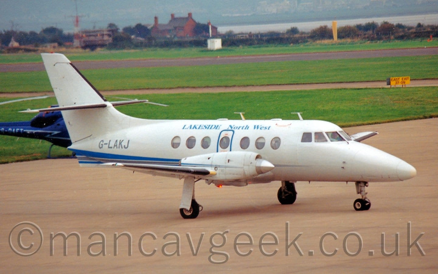 Side view of a twin propellor-engined airliner taxing from left to right. The plane is mostly white, with a blue stripe running along the body from behind the wings, blue "Lakeside North West" titles on the upper fuselage, and the grey registration "G-LAKJ" on the rear fuselage. The planes propellors are little more than a blur, as it makes it's way forwards. There is a metallic blue helicopter trying to hide behind this planes tail. Grey concrete apron fills the foreground, with acres of grass in the background leading up to a river in the distance.