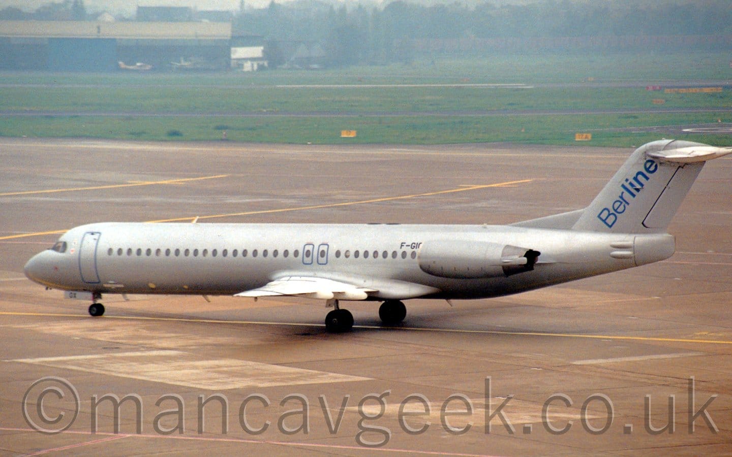 Side view of a twin engined jet airliner taxiing from right to left. The plane is painted in a silver finish, the only markings being the black registration "F-GIOX" on the upper rear fuselage, just in front of the engines which are mounted on the rear fuselage, along with blue outlines for emergency exits, and blue diagonal "BerLine" text on the tail. Grey concrete apron fills the foreground and a good chunk of the background, with grass on the far side leading up to blue hangars and trees in the distance.