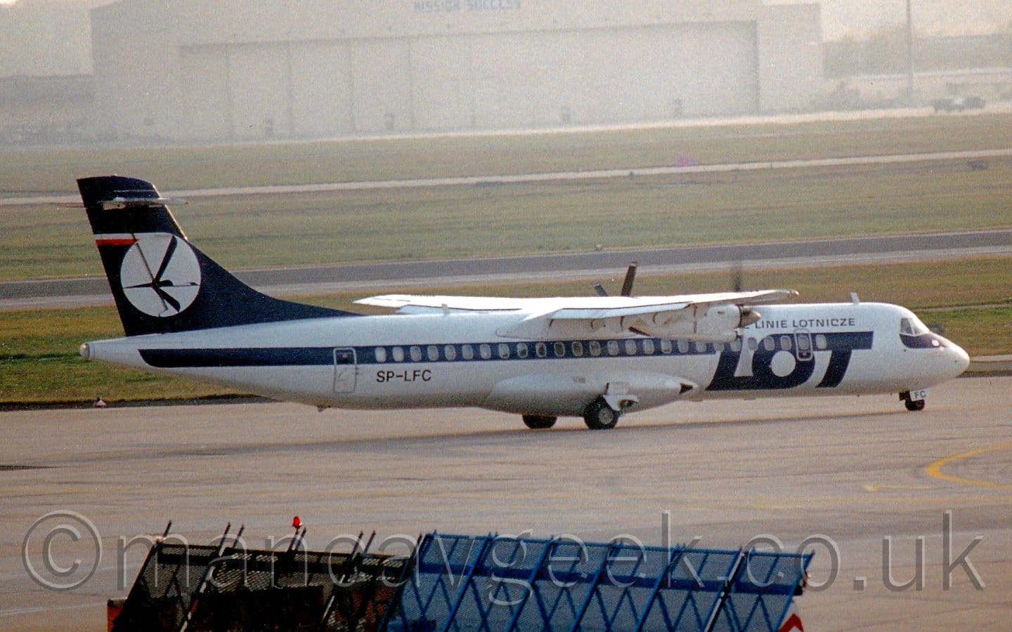Side view of a high-winged, twin propellor-engined airliner taxiing from left to right. The plane is mostly white, with a thick blue stripe running along the fuselage, covering the passenger cabin windows, and large, blue, billboard-style "LOT" titles on the forward fuselage. The tail is dark blue, with a large white circle containing a blue stylised flying bird, its wings outstretched and swept forward. Grey concrete apron fills the foreground, with a blue metal frame at the bottom middle, while grass in the background leads up to a large, cream-coloured hangar that is slowly being swallowed up by mist.