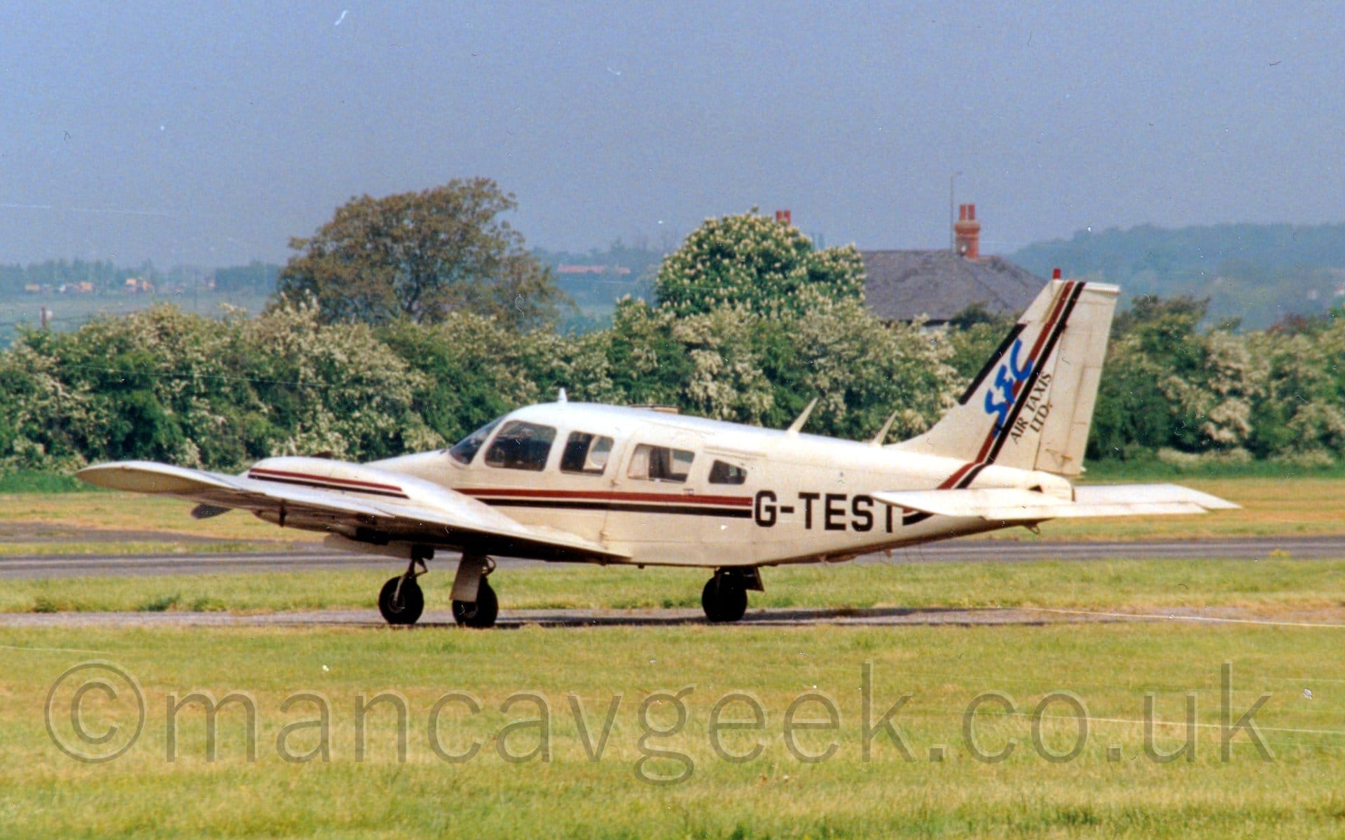 Side view of a twin propellor-engined light aircraft taxiing from right to left along a narrow grey tarmac taxiway. The plane is a light sandy-brown, with a red and black stripe running along the body and up into the tail, as well as along the engine cowlings mounted on the wings. The black registration "G-TEST" is overlaid on the stripes on the rear fuselage, with large blue text "SFC" and black text "Air taxis LTD" over the stripe on the tail. Green grass fills the foreground, with a line of trees in the background, in front of a house with a grey roof. Grey skies over trees in the distance fill the rest of the frame.