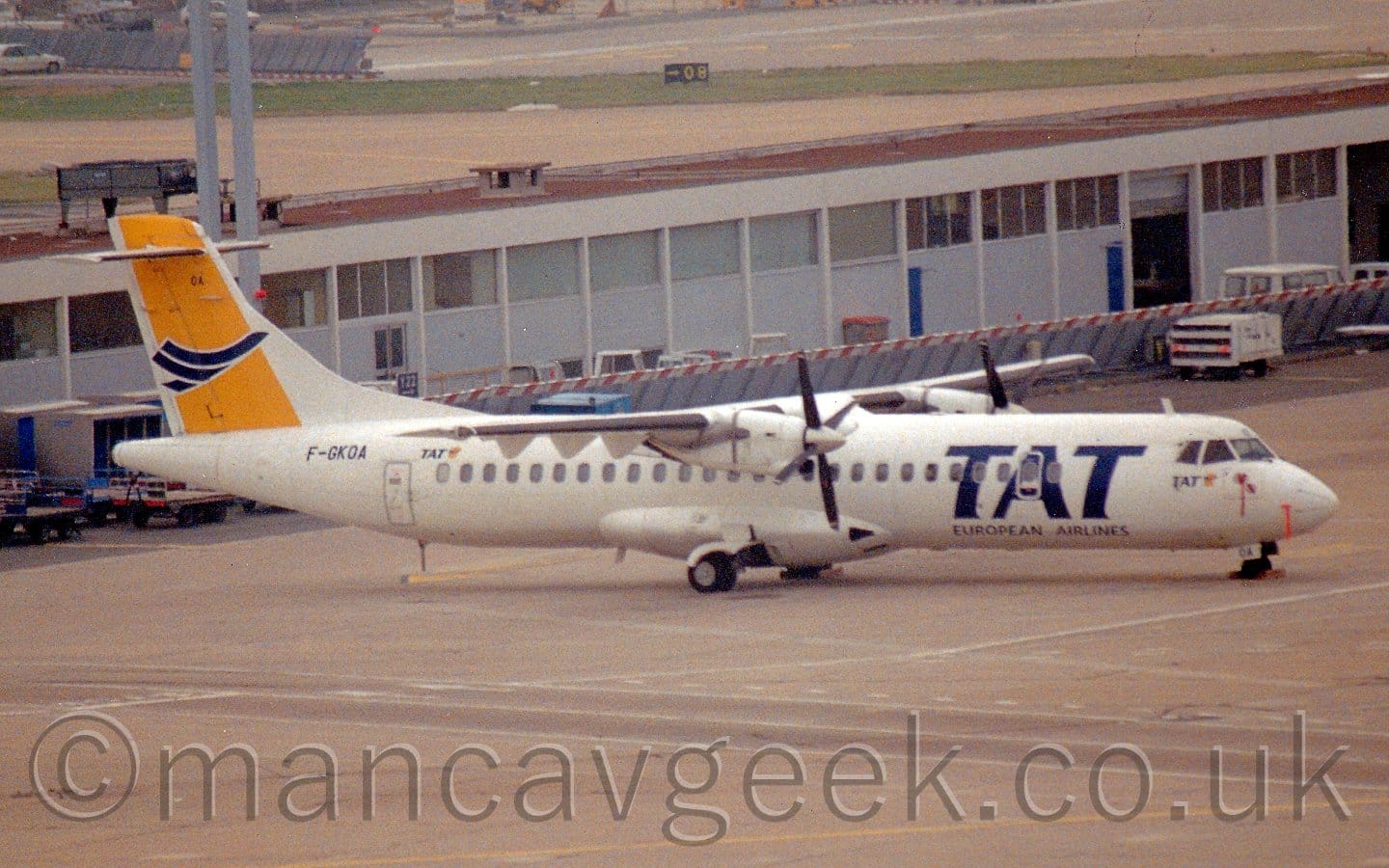Side view of a high-winged, twin propellor-engined airliner parked facing to the right. The plane is mostly white, with large, billboard-style blue "TAT" titles on the forward fuselage, with smaller blue "European Airlines" titles just below. The tail has a diagonal yellow band overlaid a stylised image of a blue flying bird, with the blue registration "F-GKOA" on the upper rear fuselage just below. Grey concrete apron fills the foreground, with a grey building with a flat red roof stretching across the background.