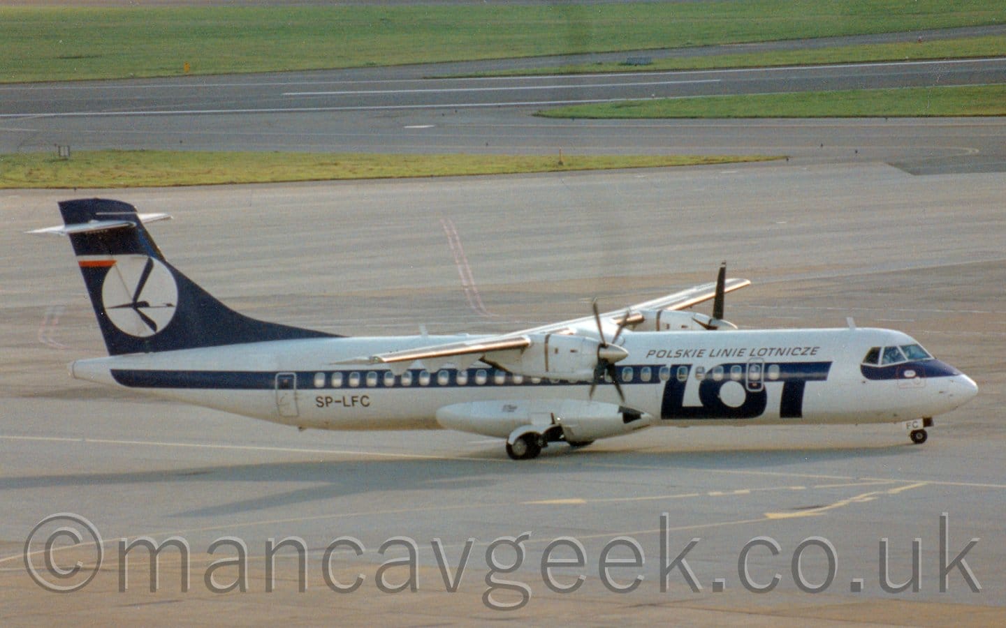 Side view of a high-winged, twin propellor-engined airliner taxiing from left to right. The plane is mostly white, with large blue letters "LOT" on the lower forward fuselage, and a blue stripe running along the body, covering the passenger cabin windows. There is some smaller, blue text in Polish on the upper forward fuselage, roughly translating to "Polish Air Lines" The tail is the same dark blue, with a white circle containing a stylised blue flying bird, it's wings outstretched and swept forwards. Grey concrete apron fills most of the rest of the frame, with grass either side of a runway in the distance.
