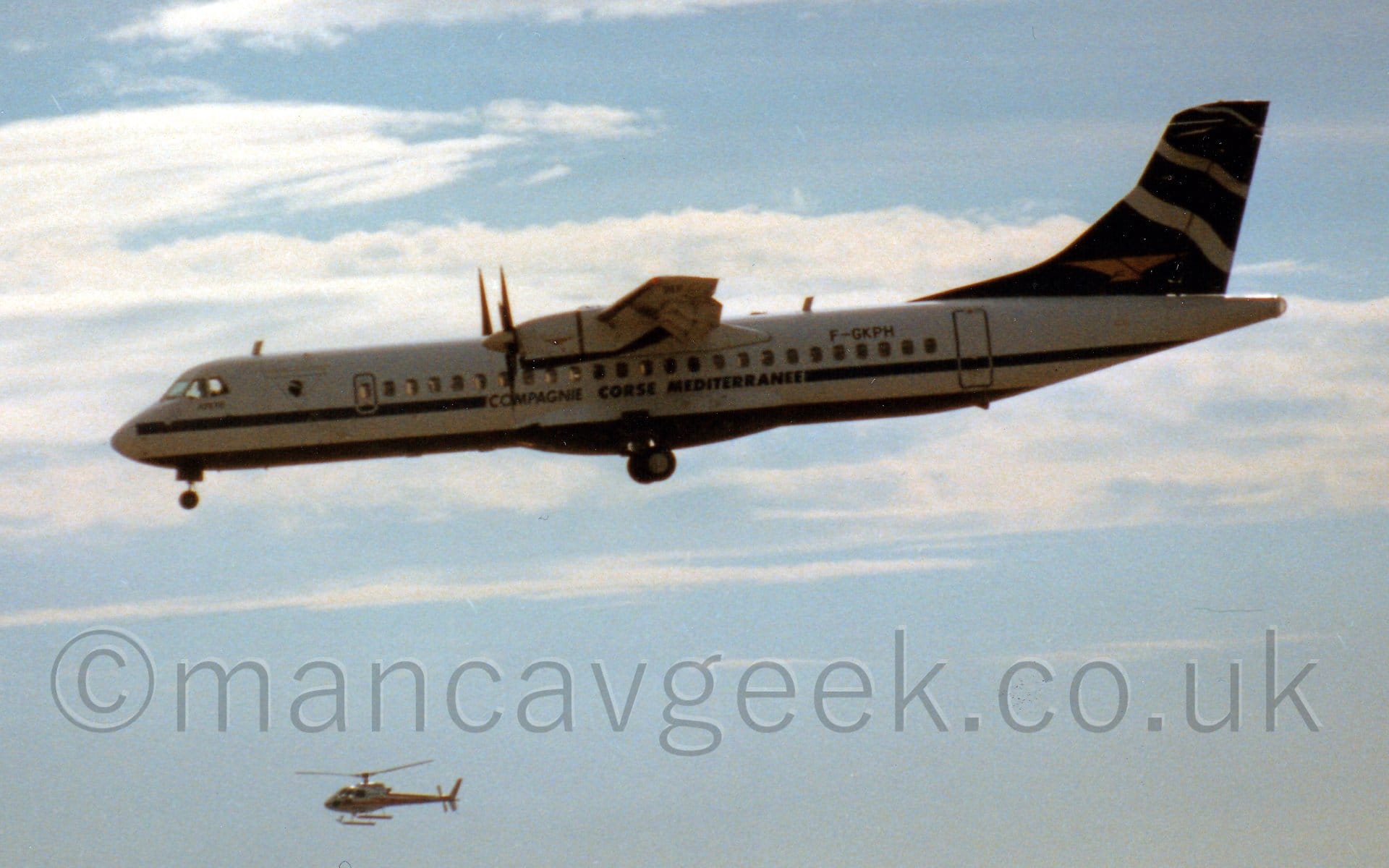 Somewhat grainy shot of a high-winged, twin propellor-engined airliner flying from right to left at a very low altitude, with it's undercarriage extended and flaps deployed from the rear of the wings, suggesting it is about to land. The plane is mostly white, with a blue belly and blue stripe running along the body just below the passenger cabin window, the latter overlaid with blue "Compagnie Corse Mediterranee" titles. The tail is dark blue, with stylised white waves and golden birds. Pale blue skies with fluffs of white cloud fill the background, with a white helicopter flying at the bottom of the frame.