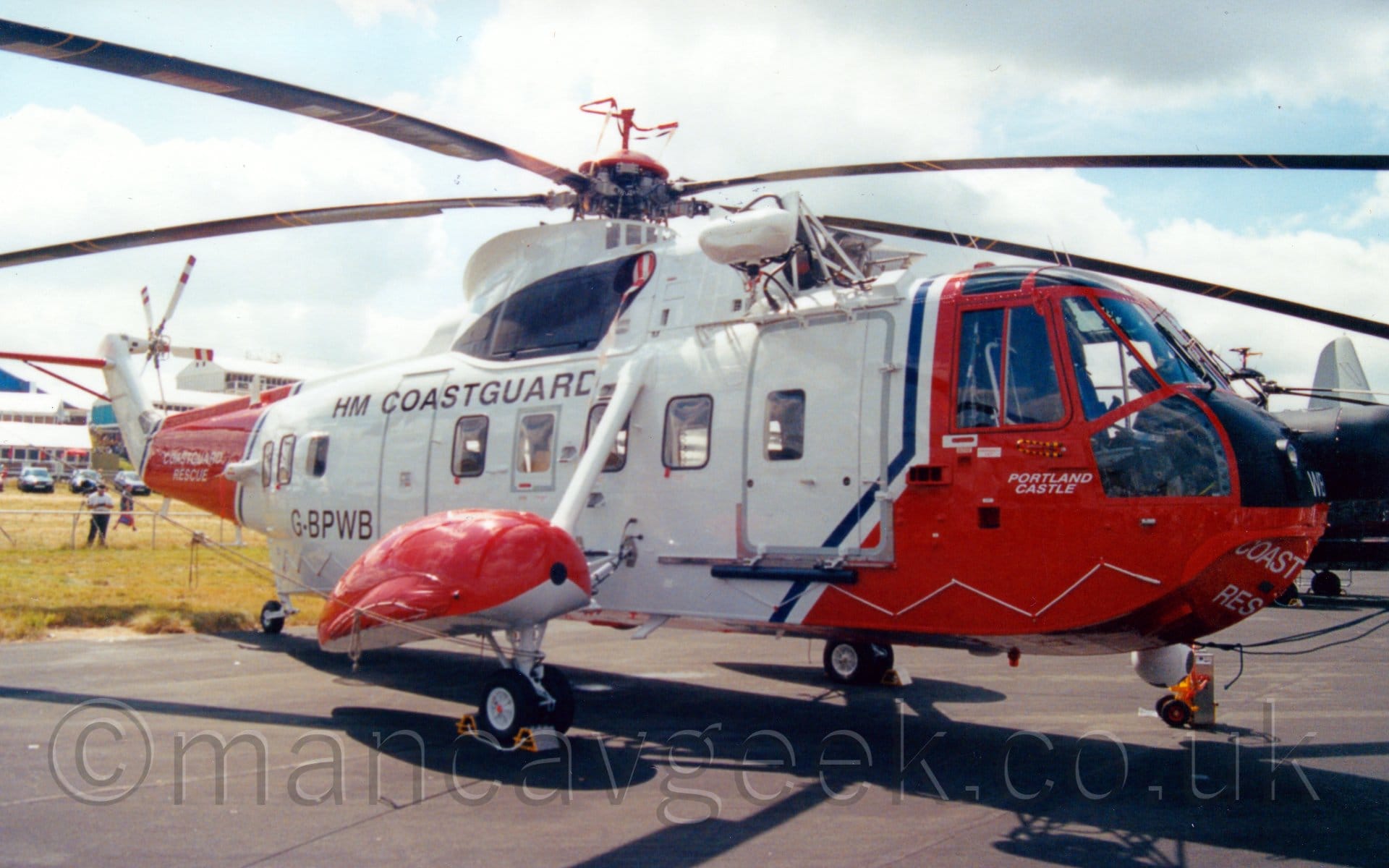 Side view of a helicopter parked on black tarmac, facing to the right. It is mostly white, with a red nose and forward fuselage, a red band around the tail-boom, and red sponsons over the main landing gear, attached to the sides of the rear fuselage. here are black "H M Coastguard" titles on the upper rear fuselage, and the black registration "G-BWPB" on the lower rear. Some other planes can be seen in the background on the right, with some buildings on the left, under bright, cloudy skies with patches of blue The helicopter is casting a strong shadow on the ground directly underneath, suggesting that the sun is almost directly overhead.