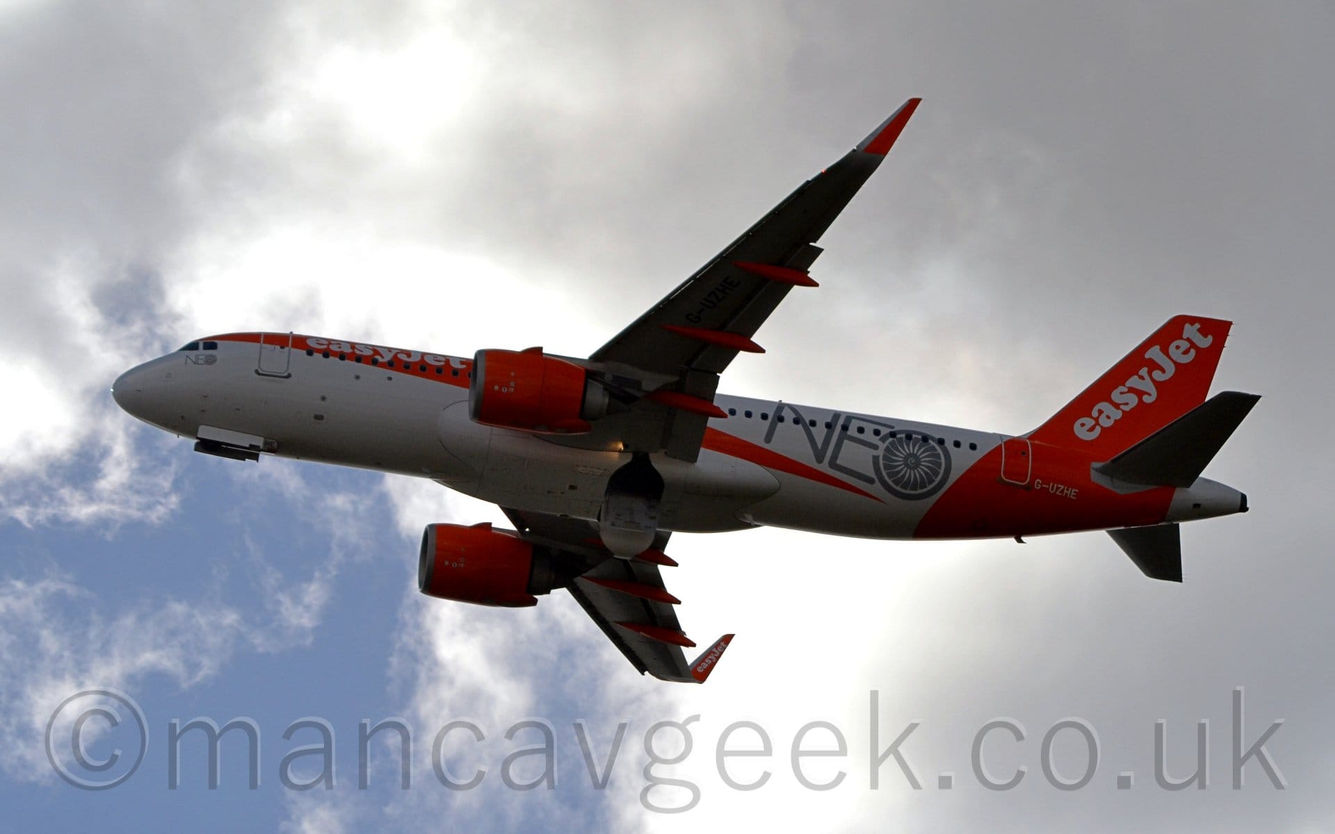 Heavily backlit photo of a twin engined airliner flying from right to left at a very low altitude, it's undercarriage in the process of being tucked away, flaps extended from the rear of the wings, and the nose raised significantly, suggesting it has just taken off. The plane is mostly white, with something that could best be described as "an orange bandana" on the upper forward fuselage, and an orange rear fuselage and tail. There are white "EasyJet" titles on the upper forward fuselage, as well as diagonally on the tail. There are additional grey "Neo" titles on the rear fuselage, the "o" being made to look like the fans at the front of a jet engine. The engines, up-turned wingtips, and the covers on the flap supports under the wings are the same bright orange. Somewhat dramatic grey skies with large bright patches, and a large blue area in the bottom left corner, fill the background.