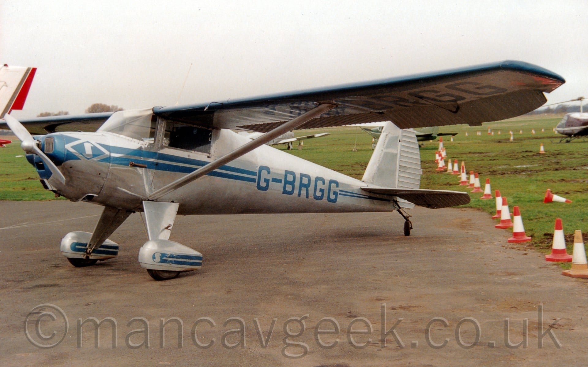 Side view of a high-winged single-engined light aircraft parked on a tarmac apron facing to the left. The plane is mostly in a bare metal finish, with a pair of blue stripes running along the body, getting narrower as they move to the rear, overlaid with the blue registration "G-BRGG" on the rear fuselage, which is repeated under the left wing. Another pair of blue stripes adorn the stripes on the spats over the main wheels. Struts support the wings, connected to the body just above the main gear legs. Grey tarmac apron fills the foreground, with a row of orange and white traffic cones separating that from muddy grass on the right of the frame. There are more light aircraft parked on grass in the background, under cloudy grey sky.