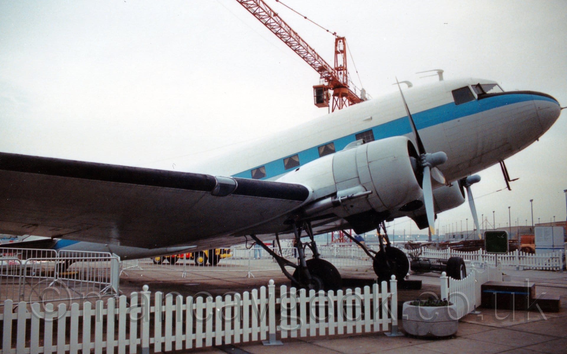 Side view of a twin propellor-engined World War 2-era airliner on display on the roof of a large building, facing to the right, behind a low white picket fence. The plane is mostly white, with a blue stripe running along the body from the nose, covering the passenger cabin windows. Bright but hazy sky fills the rest of the frame, with an orange crane looming over everything in the background.