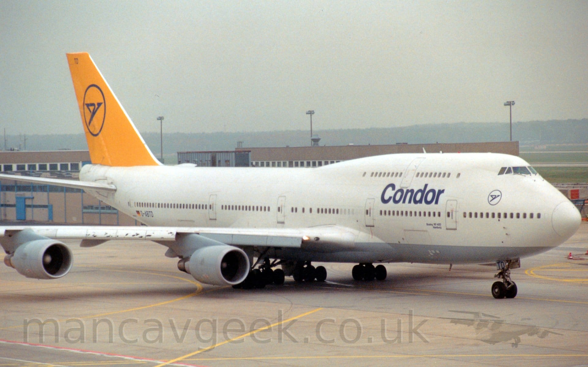 Side view of a very large, 4 engined jet airliner taxiing from left to right. The plane is mostly white, with dark blue "Condor" titles on the upper forward fuselage, between the 2 passenger decks. There is the blue outline of a circle, containing a blue stylised flying bird under the cockpit windows, a logo repeated in a larger form on the bright yellow tail. The registration "D-ABTD"  is on the lower rear fuselage, next to a German flag. Grey concrete apron fills the foreground, with a low, brown building stretching across the background, almost hidden by this behemoth of an aeroplane. Trees line the horizon, slowly fading into the hazy grey sky.