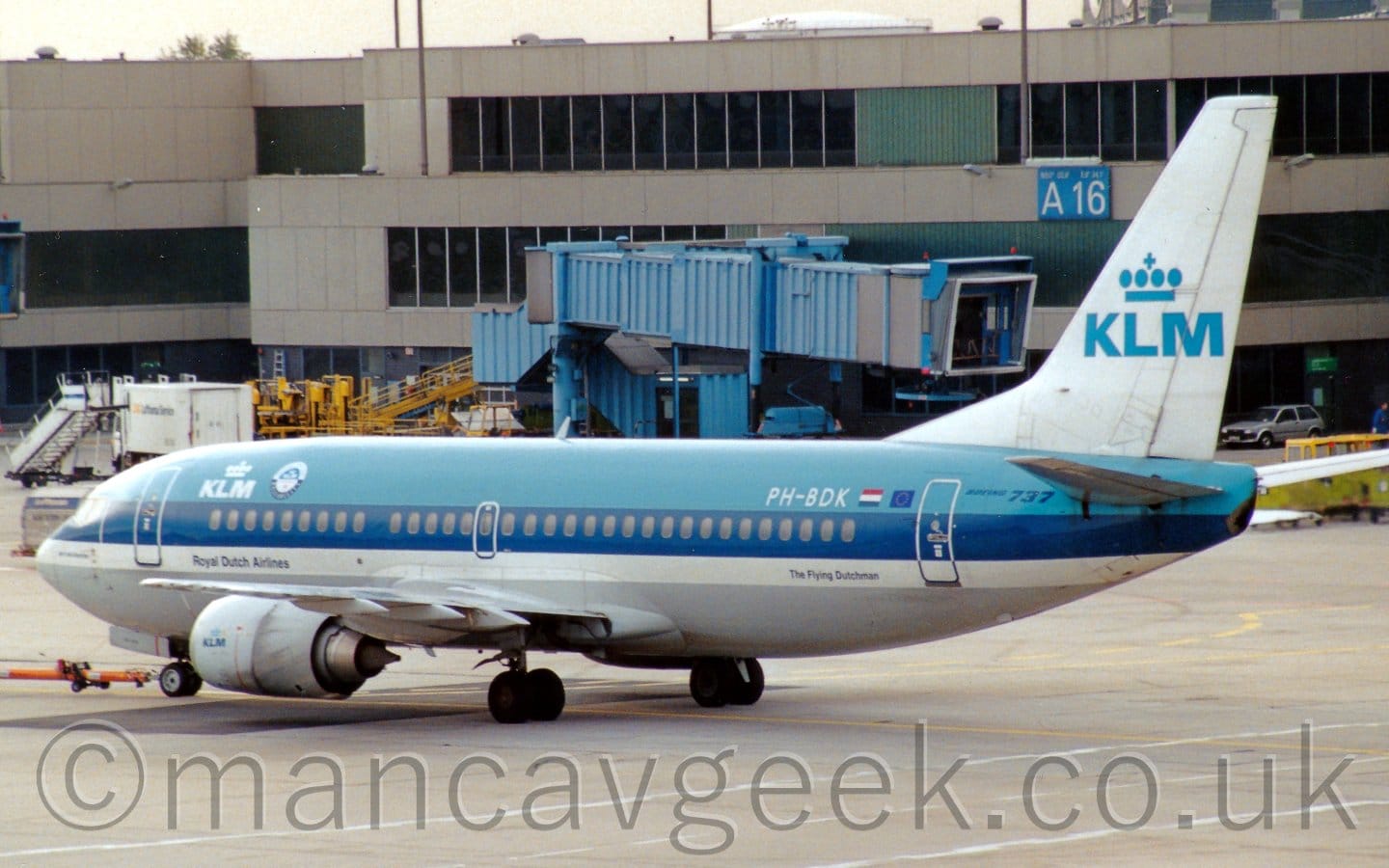Side view of a twin-engined jet airliner facing to the left but being pushed back to the right by an out-of-frame tug attached to the nose-wheel by a long orange pole. The plane is light blue on top, with a dark blue stripe running along the body covering the passenger cabin windows, a thinner white stripe below that, and a grey belly. There are white "KLM" titles under a stylised white crown on the upper forward fuselage, with blue "Royal Dutch Airlines" on the white stripe on the forward fuselage. The white registration "PH-BDK" is on the upper rear fuselage next to small Dutch and EU flags. The tail is white, with large blue "KLM" titles under a stylised blue crown. A smaller version of this logo appears on the rather dirty, white engine pods slung under the wings. Grey concrete Apron fills the foreground, while a grey terminal building stretches across the background, with a blue air bridge hanging off the side. Bright but hazy sky fills the top of the frame.