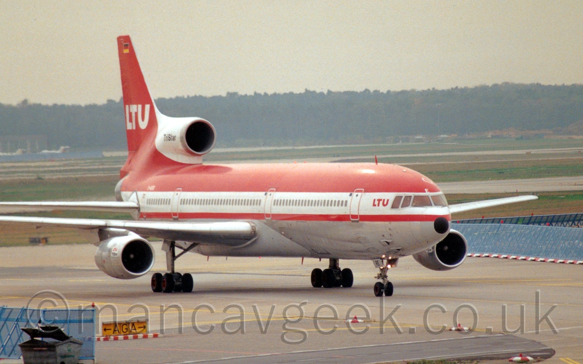 Side view of a 3 engined jet airliner taxiing from left to right. The plane is largely red, with a grey belly, and a thick white stripe running along the body, covering the passenger cabin windows, with small red "LTU" titles just aft of the cockpit windows. The tail is largely red, with white "LTU" titles, while the centre engine intake is white, with black "TriStar" text. Grey concrete taxiways fill the foreground, with wide grassed areas lining more taxiways and runways in the background, running up to trees in the distance, under bright but hazy skies.