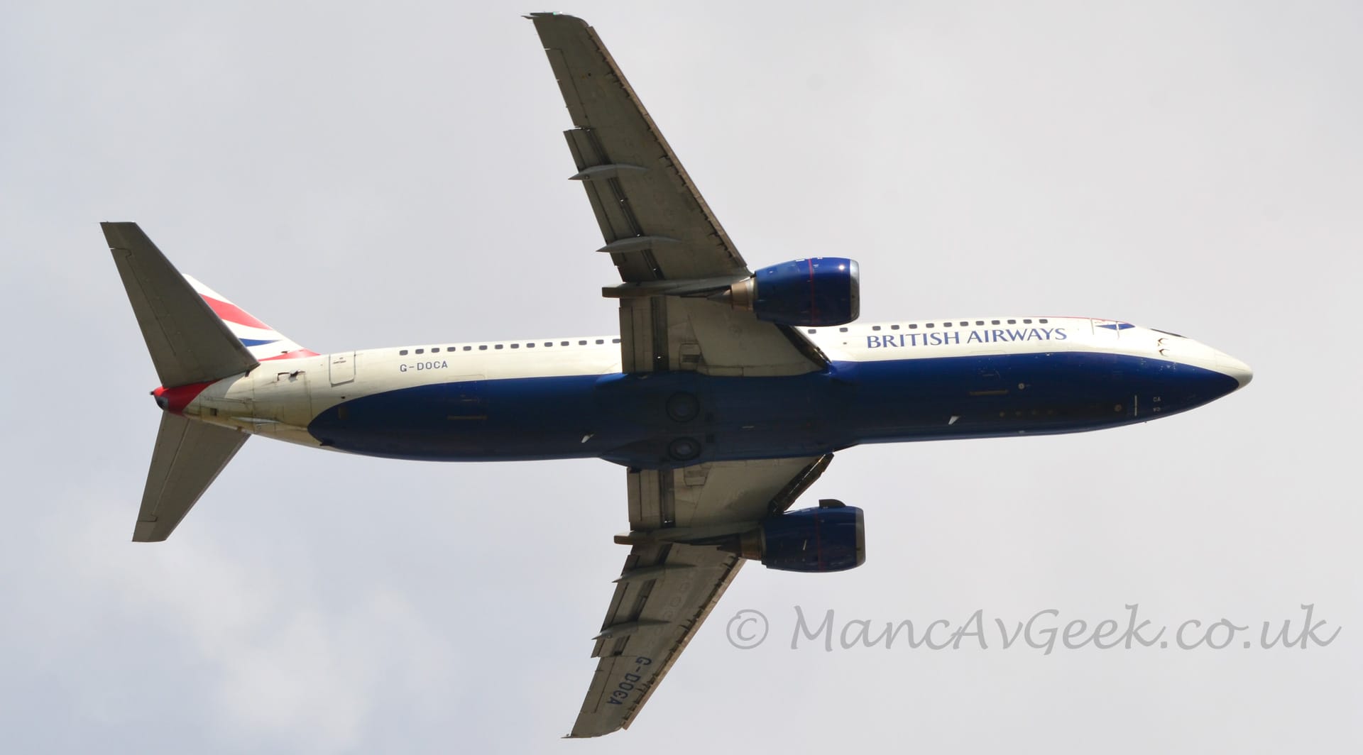 View from almost directly below a twin engined jet airliner flying from left to right with it's undercarriage already tucked away, flaps extended from the rear of the wings, and the nose raised, suggesting it has just taken off. The plane is mostly white, with a deep blue belly, and blue "British Airways" titles on the lower forward fuselage. The black registration "G-DOCA" is on the lower rear fuselage, while the white tail has wavy red and blue stripes. The engine pods under the wings are the same shade of blue as the belly. Flat grey sky fills the background.