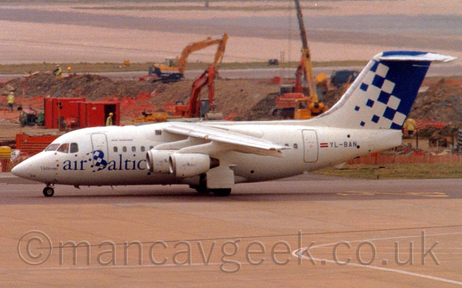 Side view of a high-winged, 4 jet engined jet airliner taxiing from right to left. The plane is mostly white, with blue "Air Baltic" titles on the lower forward fuselage, and the blue registration "YL-BAN" on the rear fuselage, next to a small Latvian flag (red field with a white stripe through the middle). The front of the tail is white, slowly turning to blue at the rear. Grey concrete apron fills the foreground, with a building site stretching across the background, with the start of a runway behind that.