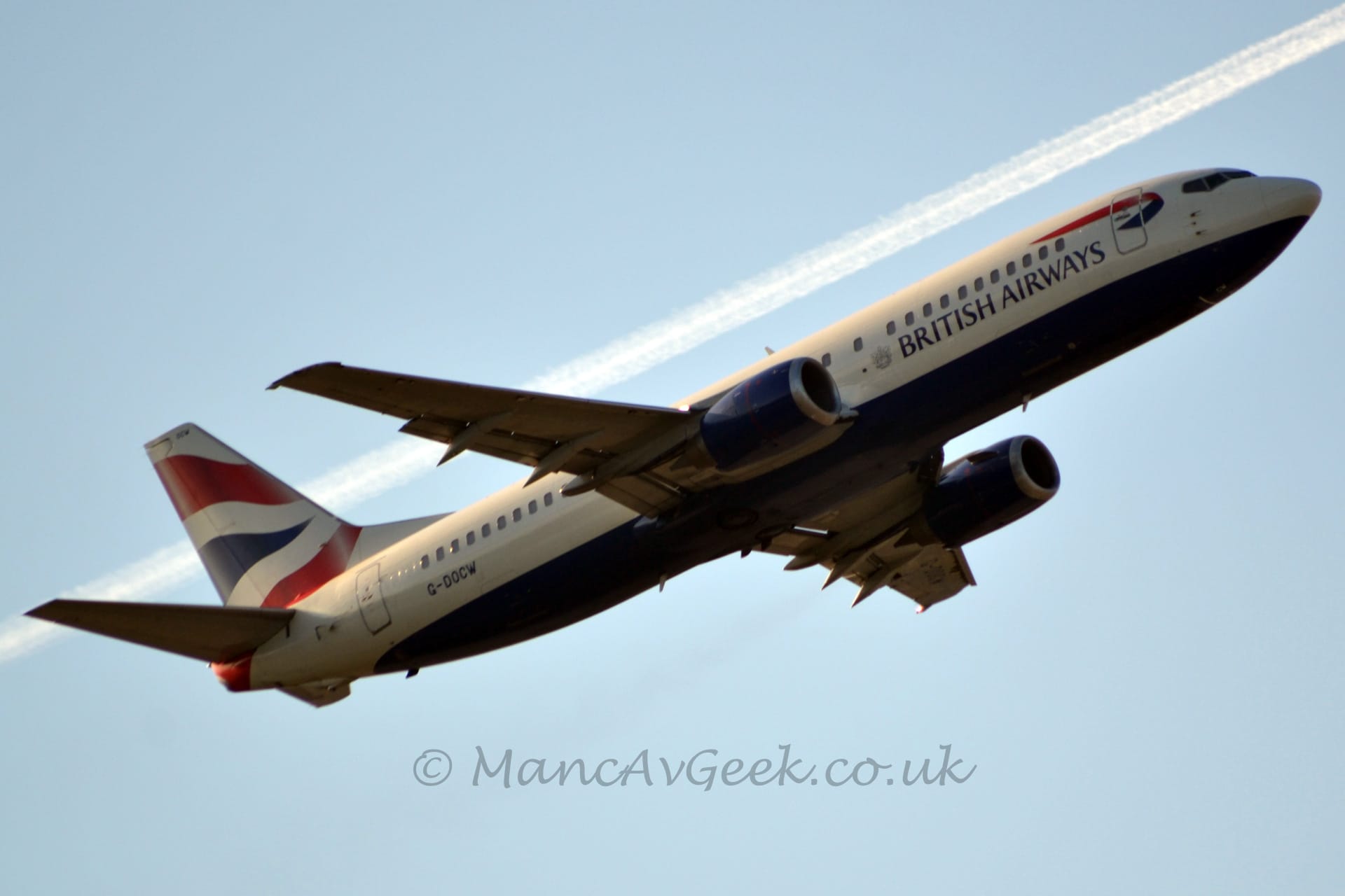 Low side view of a twin engined jet airliner flying from left to right at a very low altitude, with its undercarriage already tucked away, flaps extended from the rear of the wings, and the nose raised sharply, suggesting it has just taken off. The plane is mostly white, with a dark blue belly and dark blue "British Airways" titles on the lower forward fuselage, under a twisted blue and red ribbon. There are wavy red and blue stripes on the tail and the black registration "G-DOCW" on the lower rear fuselage. The engine pods, mounted under the wings, are the same dark blue. Blue skies fill the rest of the frame, with the contrail of a passing high-level jet airliner passing through the middle of the frame, heading in pretty much the same direction.