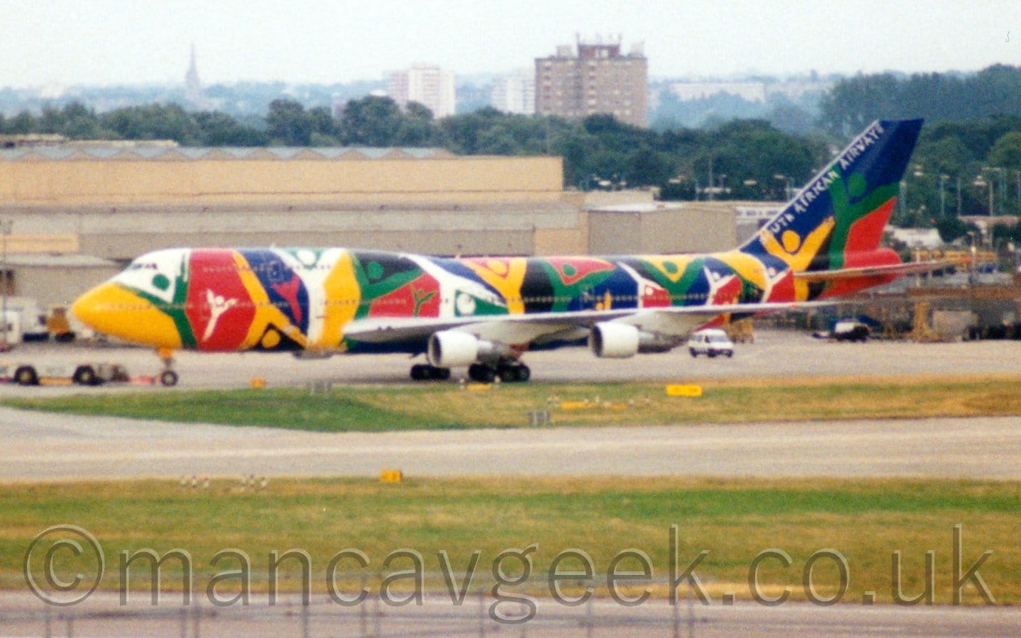 Slightly grainy shot of a very colourful, very large 4 engined jet airliner being towed from right to left by a low-slung grey tug attached to the nose-wheel by a long pole. The plane is covered in seemingly random patches of colour, but closer examination reveals that most of these are stick figures with arms raised in a celebratory pose, with more colours defining the negative space between them. The tail is mostly dark blue, with white "South African Airways" tiles along the leading edge. Green grass lines several taxiways in the foreground, with a wide, sandy brown building in the background. Trees and tall residential buildings in the distance are slowly vanishing into the hazy grey sky.