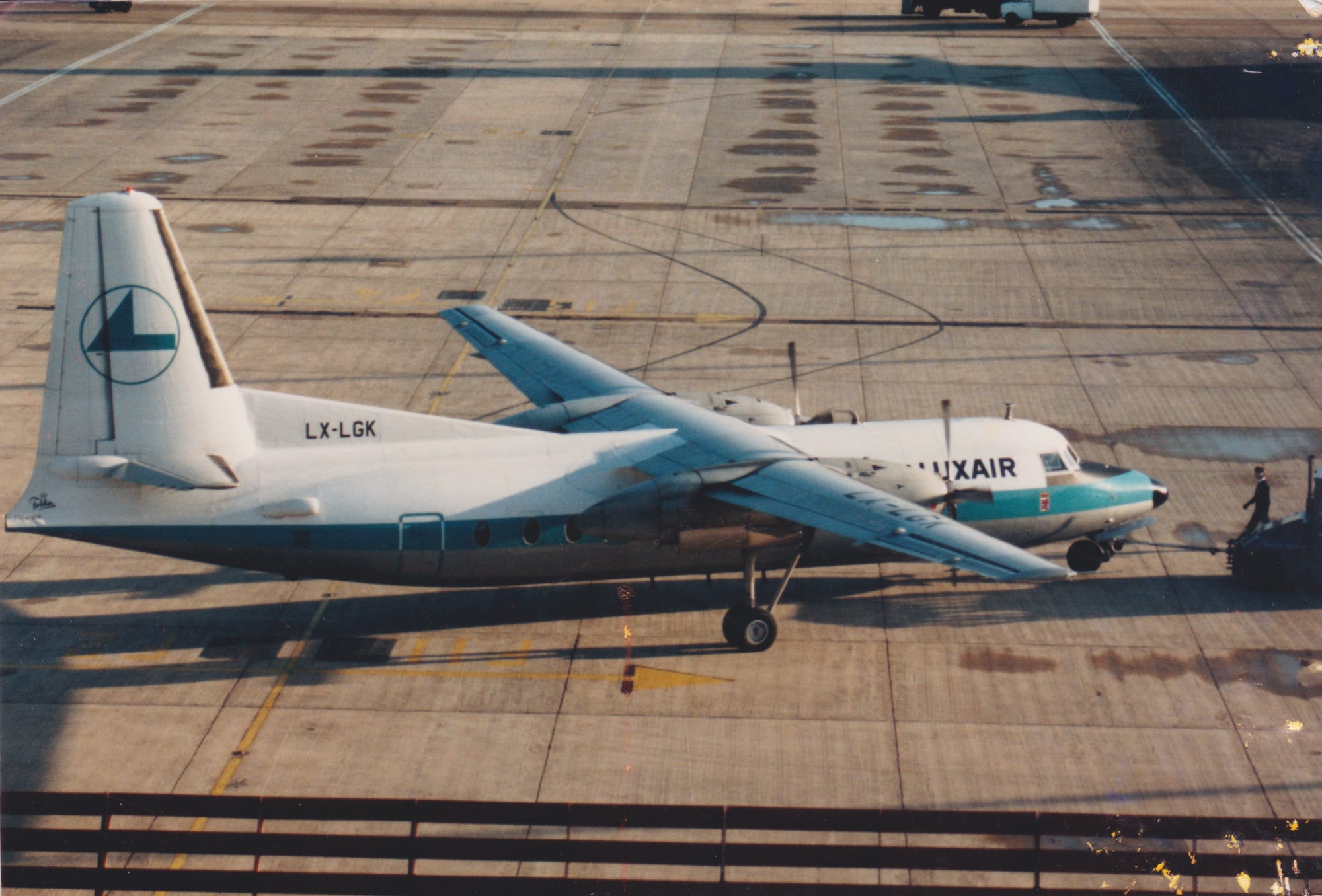 High side view of a high-winged, twin propellor-engined airliner facing to the right but being pushed back to the left by an out-of-frame tug attached to the nose-wheel by a long blue pole. The plane is mostly white, with a grey belly, and a thick, light blue stripe running along the body. There are black "LuxAir" titles on the upper forward fuselage, between the cockpit windows and the wing. The tail is white, with the blue outline of a circle containing a rather angular, blue, stylised flying bird, and the black registration "LX-LGK" on a fillet at the front. Grey concrete apron with occasional damp patches fill the rest of the frame.