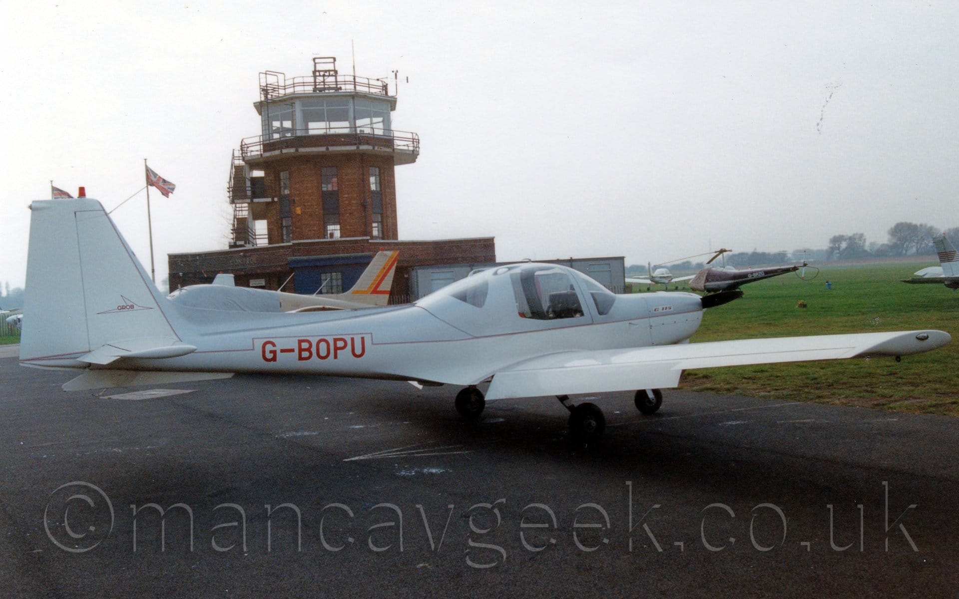 Side view of a single-engined light aircraft parked on black tarmac facing to the right and slightly away from the camera. The plane has a grey, almost metallic upper body and white lower body and tail, with a thin red pin-stripe separating the 2 colours. Ther red registration "G-BOPU" is on the rear fuselage, a white box around it cutting into the grey area. Several other planes are parked in the background, mostly on grass on the right of the frame, but also one on tarmac on the left. A red brick control tower with a heavily glazed top floor dominates the background on the left, reaching into the grey sky.