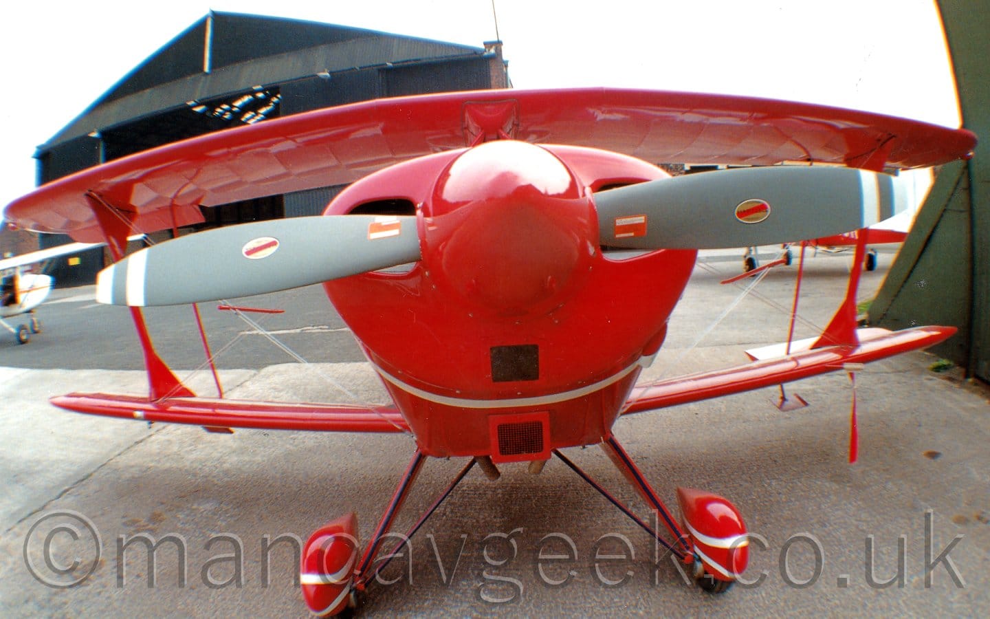 Close-up head-on shot (with a VERY wide angle lens) of a bi-plane light aircraft parked on grey concrete facing the camera. The plane is almost entirely red, with white stripes around the engine cowling, and around the spats covering the main wheels, as well as a 2 bladed grey propellor. The prop has been arranged so it is horizontal, quite possibly for any passing geek who just happens to be taking photos. The corner of a green hangar can be seen on the right of the frame, with a larger black hangar with it's doors wide open can be seen in the background on the left, with a small white plane parked in front of it. Flat white sky fills the rest of the frame.
