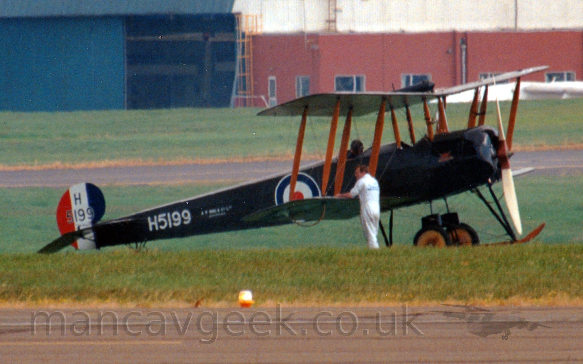 Side view of a single-engined bi-plane trainer aircraft parked facing to the right on grass. The plane has a dark brown, almost black body, with a pair of open cockpits, and a blue, white, and red roundel just aft of the rear cockpit. Red struts connect the wings, the top wing suspended over the forward fuselage. The white serial "H5199" s painted on the rear fuselage, and repeated on the small, curved tail which has red, white and blue vertical bars. A man s standing in front of the plane, holding onto the leading edge of the lower right wing. A black runway s at the bottom of the image, but it looks as if the plane is going to take off from the grass. A red and white building next to a blue hangar fills the background.