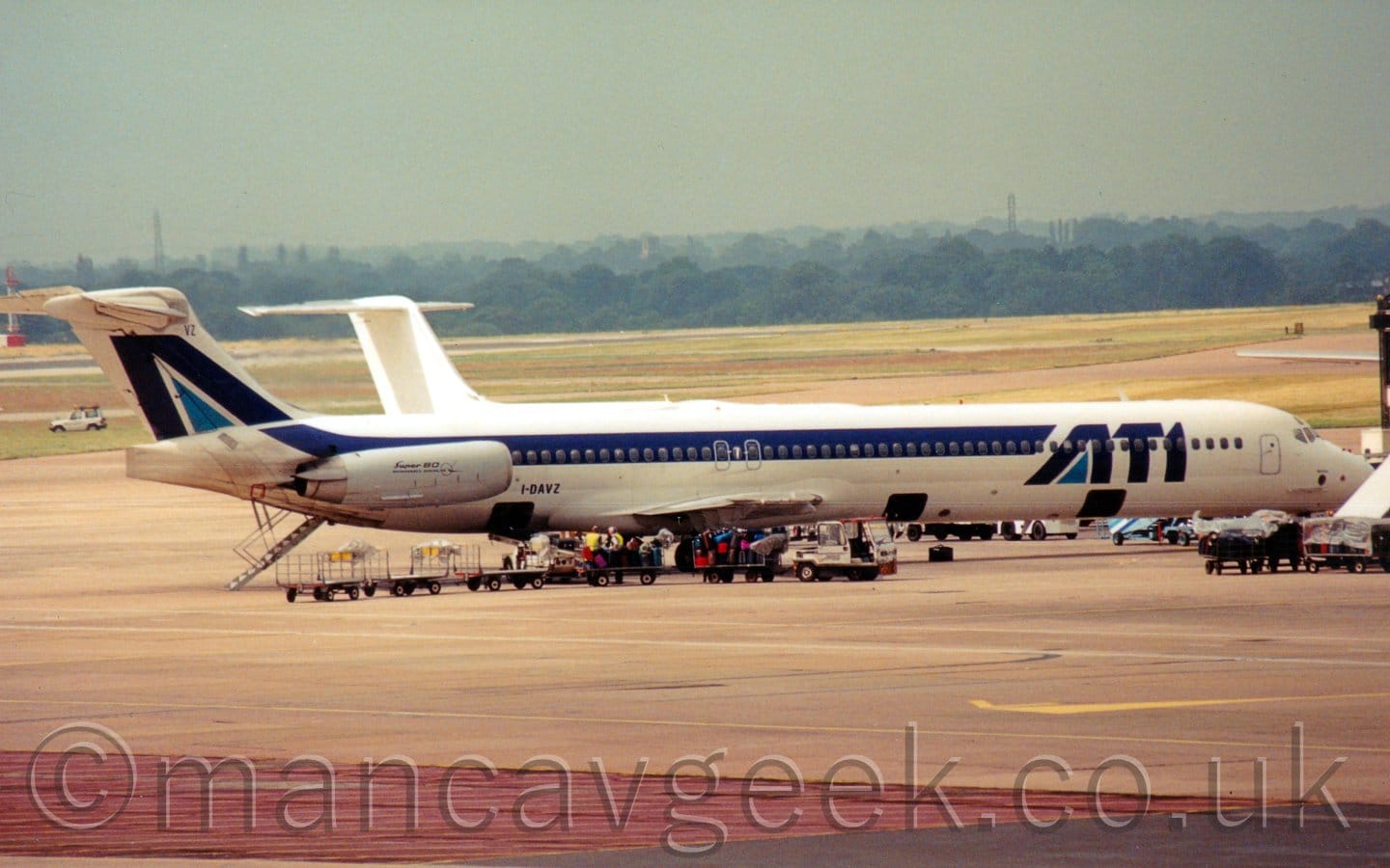 Side view of a twin-engined jet airliner parked facing to the right. The plane is mostly white, with blue "ATI" titles on the forward fuselage, and a dark blue stripe running along the body, covering the passenger cabin windows. The tail has a dark blue chevron with a light blue triangle in the middle. The engines mounted on the sides of the rear fuselage are grey, with "Super 80" text in black. Grey concrete apron fills the foreground, with a strip of red right at the bottom, while grass in the background runs out to trees in the distance. The tail of a white jet airliner can be seen immediately behind this plane. Grey sky fills the rest of the frame.