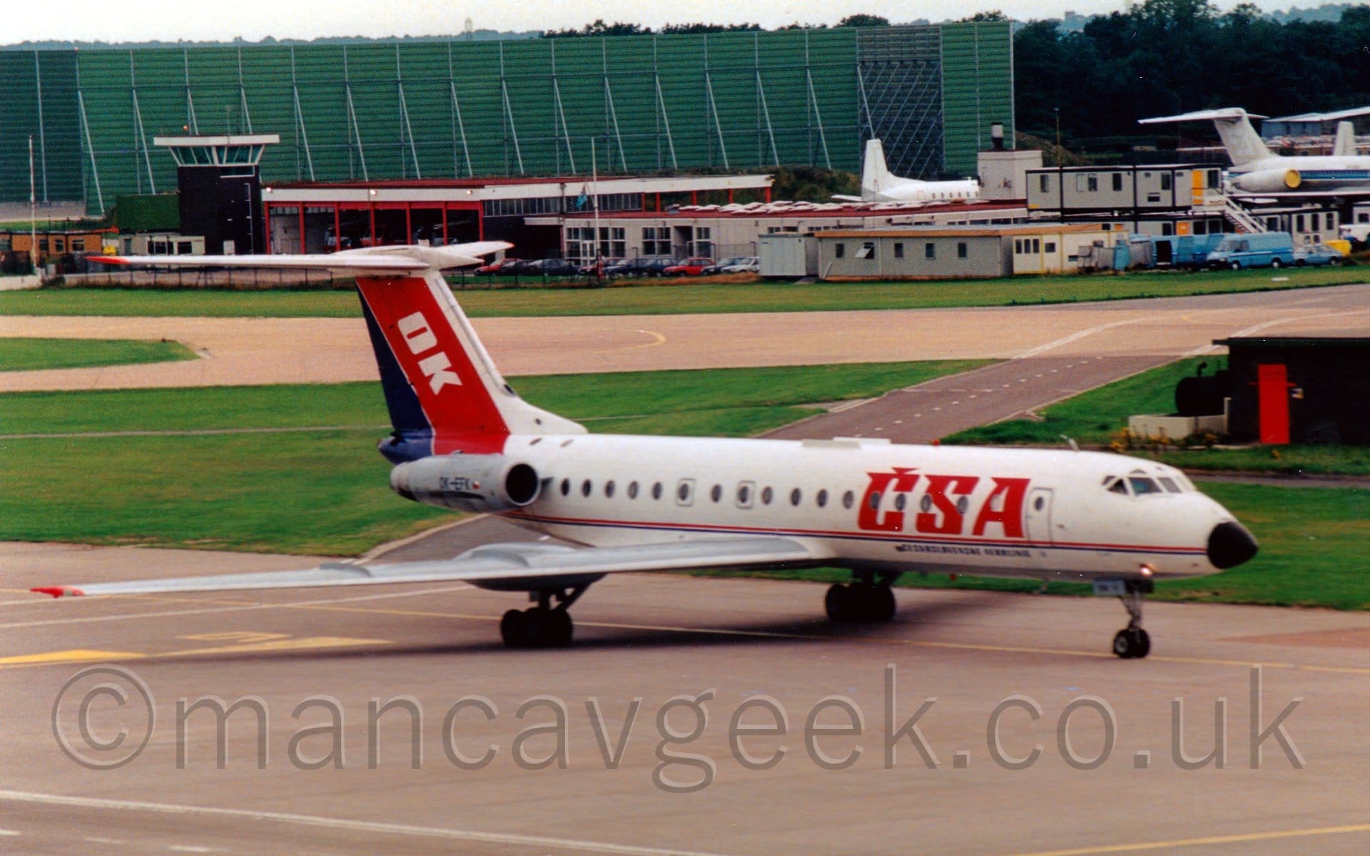 Side view of a white, twin engined jet airliner taxiing from left to right. The planes engines are mounted on the sides of the rear fuselage, under the red and blue tail. There are large red "CSA" titles on the forward fuselage, above smaller blue text in Czech that translates to "CzechSlovak Airlines". A thin blue and red stripe runs along the body from the black nose-cone. The tail is red and blue, with white letters "OK" on the red part. The main landing gear is in a strange position, at least compared to more modern airliners, attached to pods at the rear of the wing, about 1/3rd of the wingspan out from the body. A grey taxiway fills the foreground, separated from another taxiway by a wide strip of grass in the background. Various buildings, mostly temporary, pre-fabricated buildings, stretch across the background, with a large, green, box-like metal frame on the left behind the airport fire station, while the tails of several other airliners can be seen in the top right corner., under bright but hazy sky.