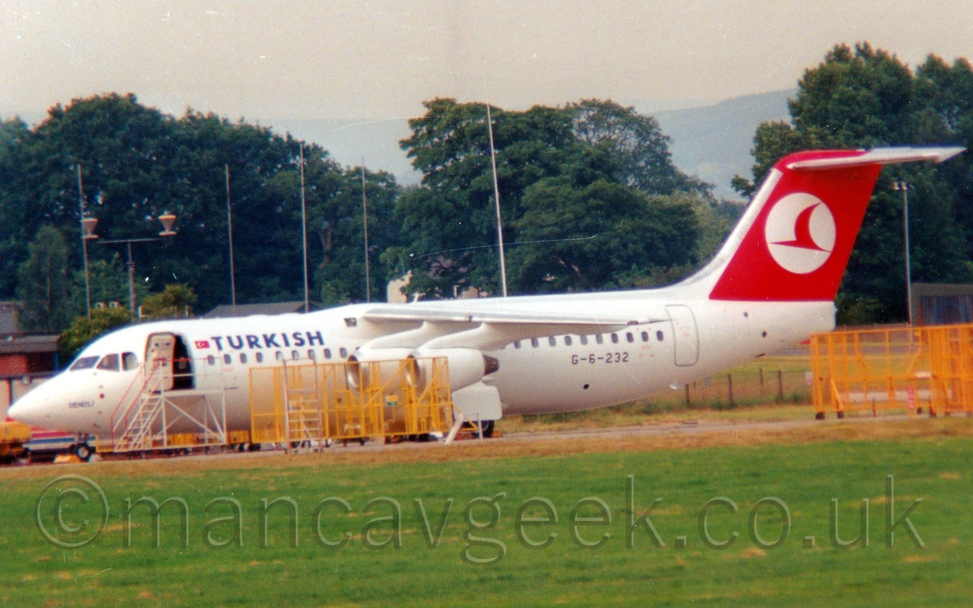 Side view of a high-winged, 4 engined jet airliner parked facing to the left. The plane is mostly white, with blue "Turkish" titles on the upper forward fuselage, next to a small Turkish flag. The black registration "G-6-232" is on the lower rear fuselage. The tail is red, with a white circle containing a stylised red flying bird. There are several yellow metal frames parked between the camera and the plane, partially obscuring the forward fuselage, as well as some on the right of the image, and a white set of airstairs parked next to the open forward door. Green grass fills the foreground, with trees lining the background, under grey sky.