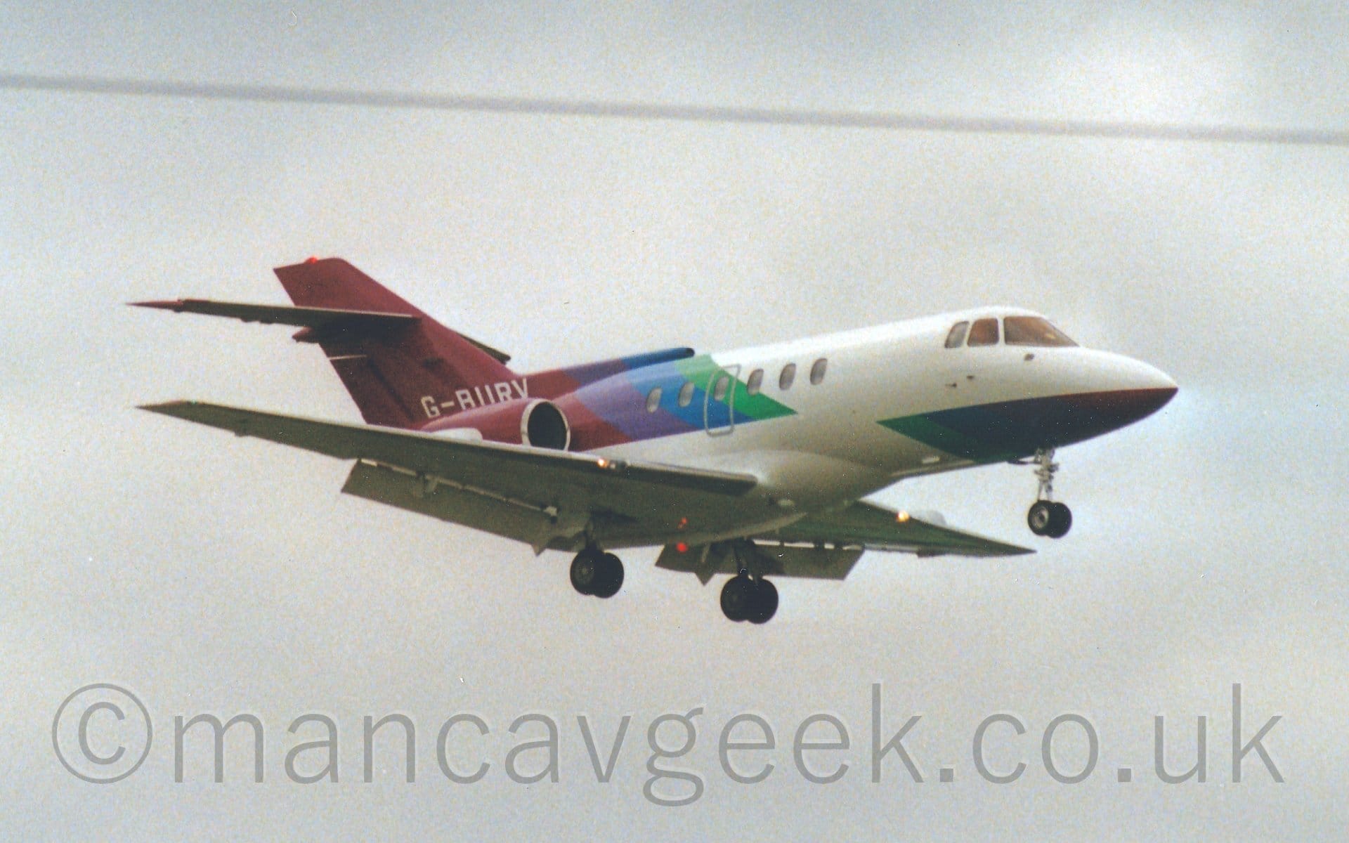 Side view of a twin engined BizJet flying from left to right at a low altitude with flaps extended behind the wings and the undercarriage lowered, suggesting that the plane is about to land. Coudy grey sky fills the rest of the frame.