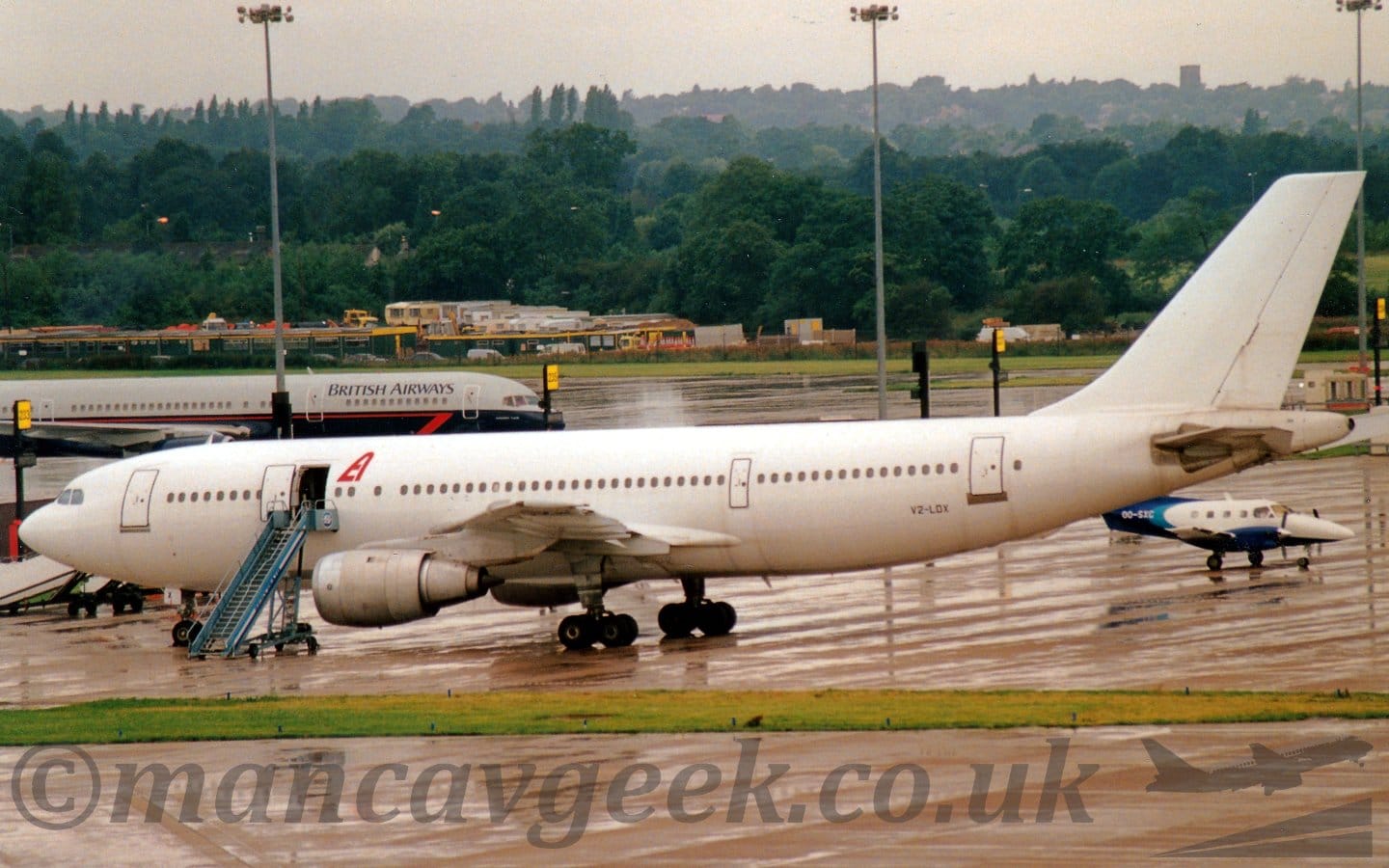 Side view of a twin engined jet airliner in a "Polar bear in a blizzard" livery, parked facing to the left. The only markings are a large red letter "A" next to the open forward mid-cabin door, and the black registration "V2-LDX" on the lower rear fuselage, jut forward of the rear cabin door. There is a set of blue mobile air stairs next to the open cabin door. A grey taxiway stretches across the frame, with a strip of grass separating that from the apron area this plane is parked on. A couple of other planes are visible in the background - a small white and blue plane under the tail on the right of the frame, and a grey and dark blue one on the left, both facing to the right. Trees in the distance slowly vanish into bright but hazy sky.