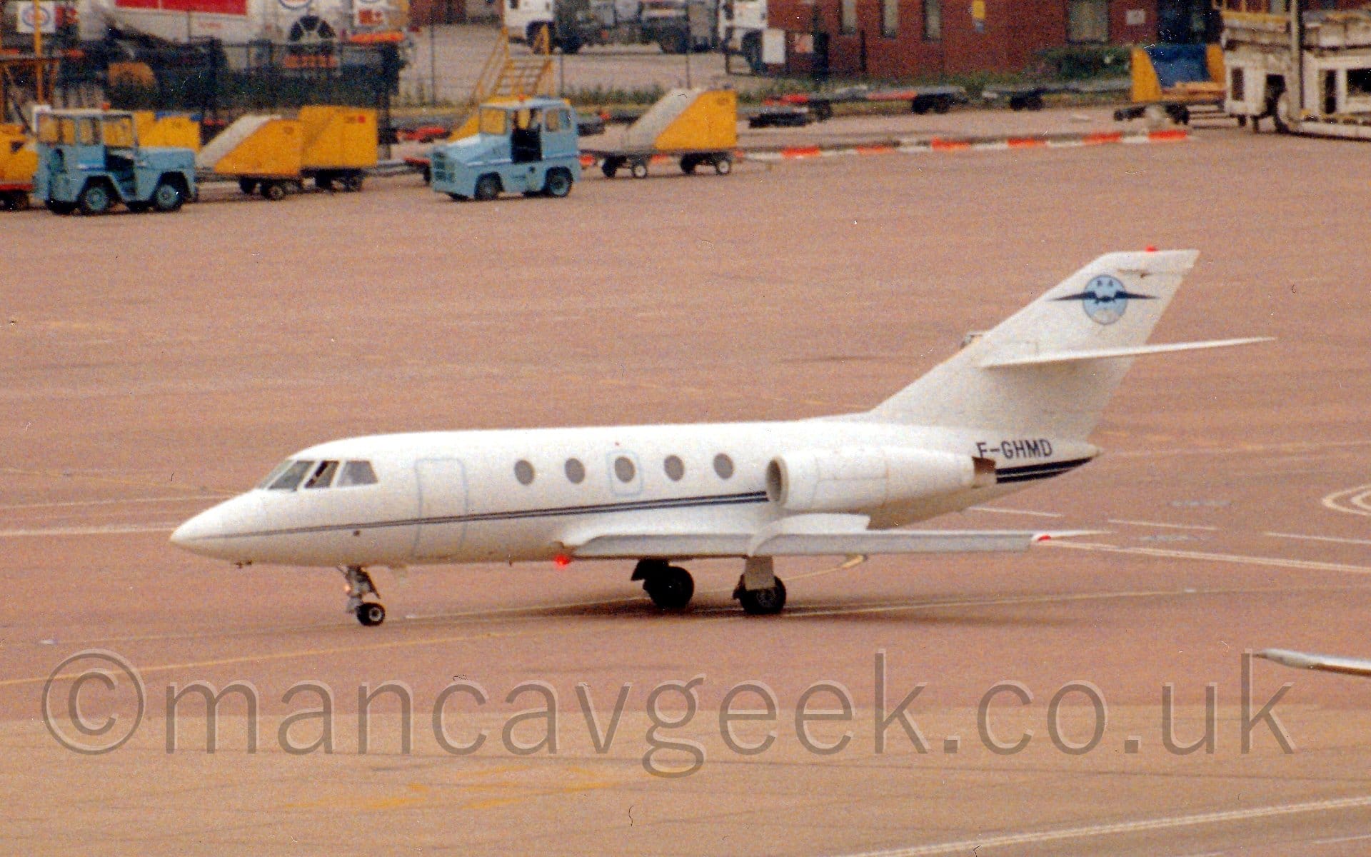 Side view of a twin engined BizJet taxiing from right to left. The plane is mostly white, with a thin blue line running along the body, getting slightly wider as it moves rearwards. The black registration "F-GHMD" is on the tail cone, under the white tail, with a circular light blue loge overlaid with dark blue wings at the top of the tail. Red-tinged concrete apron fills the foreground and most of the background, with blue trucks and yellow baggage carts parked at the top of the frame, in front of brown buildings and white fuel trucks.