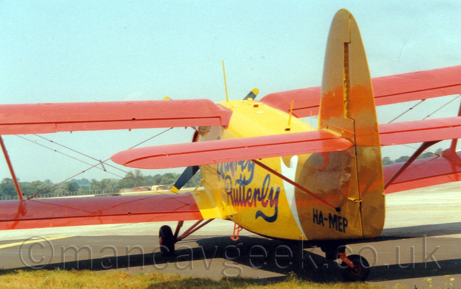 Rear view of a single-engined bi-plane airliner parked on tarmac facing away from the camera. The plane is largely yellow, with red wings and tail, and blue "Utterly Butterly" titles on the side of the rear fuselage. The black registration "HA-MEP" is on the rear fuselage, under the horizontal stabiliser. Pale blue sky fills the rest of the frame.