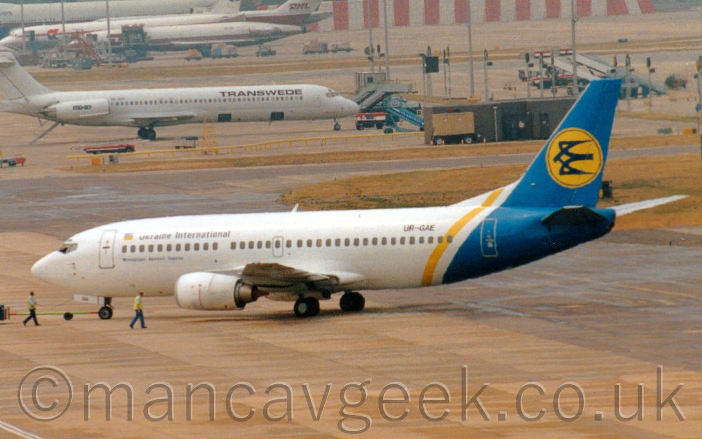 Side view of a white twin engined jet airliner facing to the left but being pushed back to the right by an out-of-frame tug attached to the nose-wheel by a long white pole. The plane is mostly white, with a blue rear fuselage and tail, with a yellow band around the rear fuselage. There are black "Ukraine International" titles on the upper forward fuselage, next to a yellow and blue Ukrainian flag, while the black registration "UR-GAE" is on the upper rear fuselage. There is a yellow circle in the middle of the blue tail, containing a stylised blue flying bird. Grey concrete apron fills the foreground, with a couple of men walking alongside the planes nose as it is being pushed backwards. More concrete apron stretches off into the distance in the background, with several white planes parked in the top left corner.