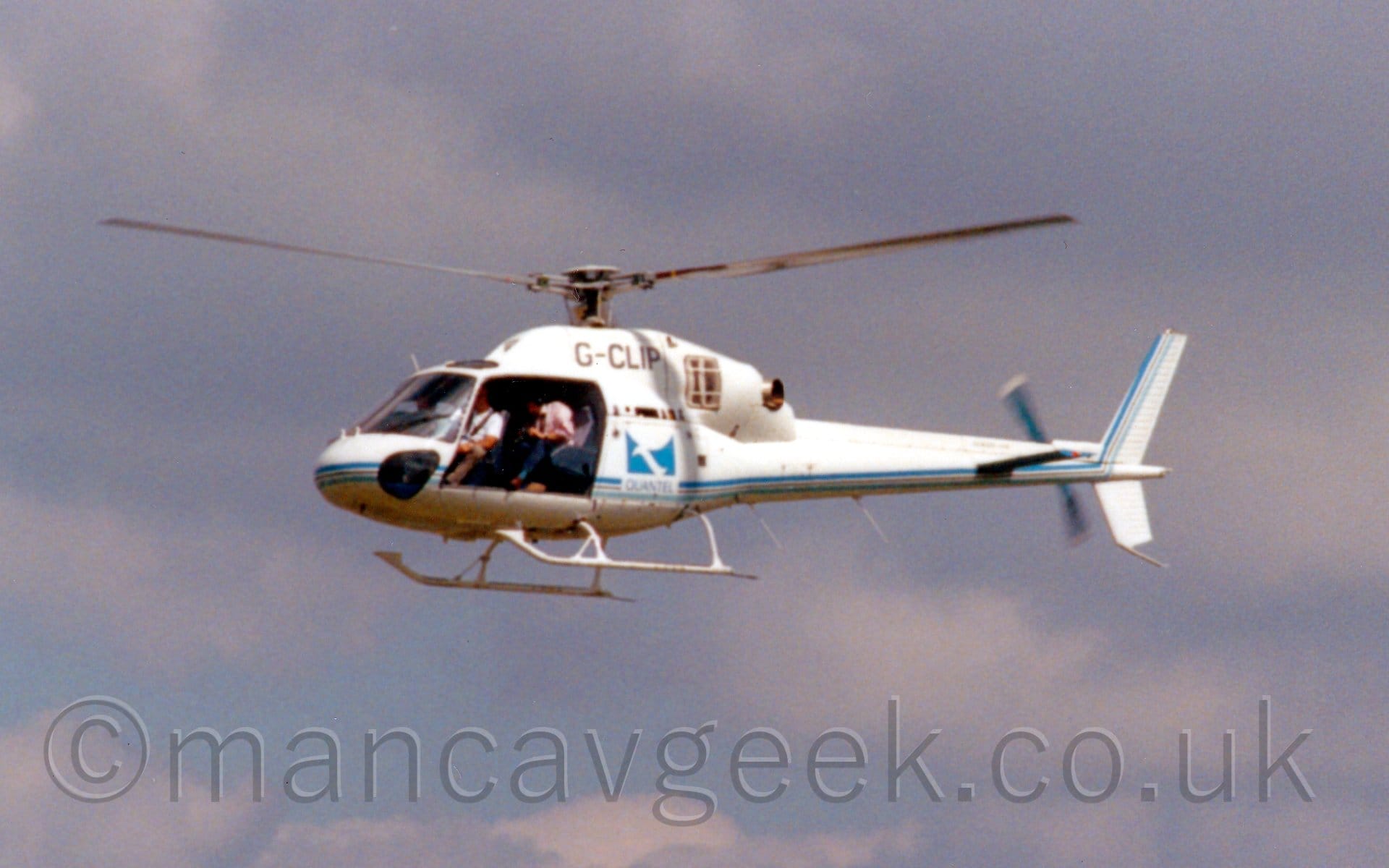Side view of a small helicopter flying from right to left at a low altitude. The chopper is mostly white, with a blue and grey stripe running along the lower body and tail boom, and a blue square with a stylised upper case white letter "Q", with the word "Quantel" at the bottom. The black registration "G-CLIP" is on the side of the engine cowling on top of the body. Blue sky with streaks of grey cloud (or is it grey cloud with streaks of blue sky? Who knows?) fill the background.