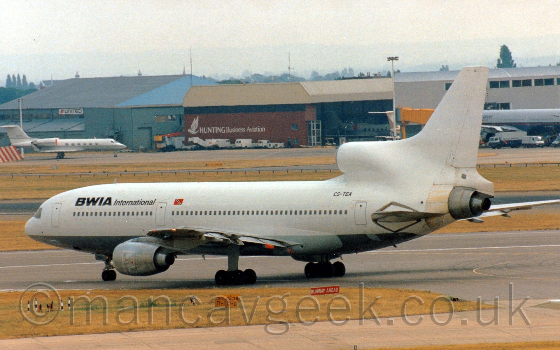 Side view of a 3 engined jet airliner taxiing from right to left on a black taxiway marked with white and yellow lines. The plane is mostly white, with a grey belly, and black BWIA International titles on the upper forward fuselage, and the black registration "CS-TEA" on the upper rear fuselage, under the centre-engine inlet. There is a lot of orange-brown staining around the rear fuselage, just in front of the exhaust outlet of the centre engine. Yellowing grass and another taxiway fill the foreground, with a grey terminal building on the right of the background, with a red and sandy brown hangar with grey "Hunting Business Aviation" titles in the middle of the frame, and a blue hangar on the left of the background. Hazy grey sky fills the rest of the frame.