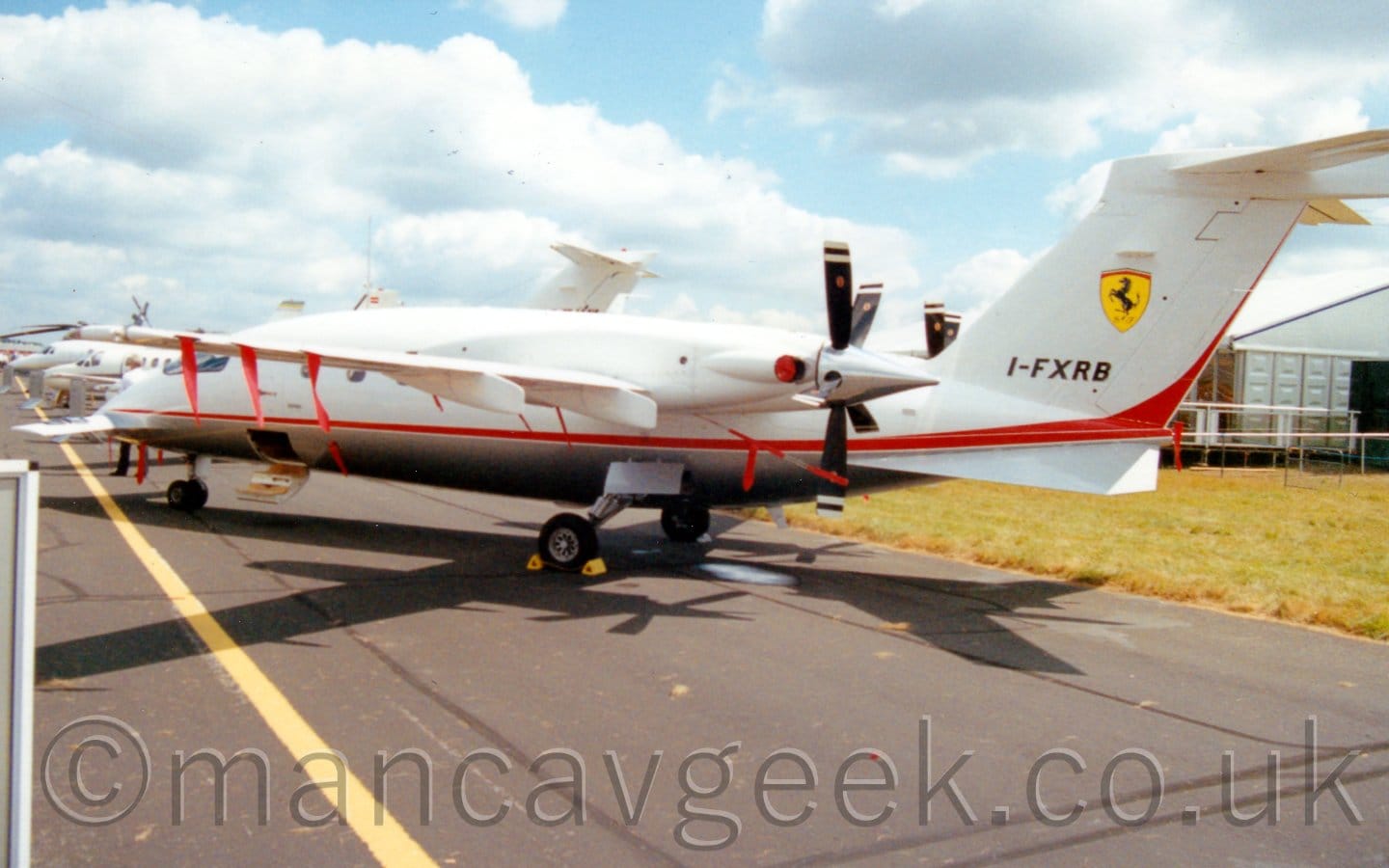Rear side view of a twin propellor-engined BizProp with the engines mounted pointing backwards of the rear of the wings. The plane is mostly white, with a grey belly, and a red stripe running along the body from the nose, before sweeping up into the rear of the tail. The black registration "I-FXRB" is on the lower part of the tail, with a yellow shield in the middle, containing an image of a prancing black horse, with a thin red and green band at the top of the shield. There are red covers on engine exhaust outlets, with red ribbons attached to several parts of the rear of the wings and on the fuselage. The bottom half of the cabin door is hanging open, forming a set of stairs, while the upper half is closed. There is a small aerodynamic lifting surface on the nose-cone. Black tarmac taxiway fills the foreground, with grass in front of a white temporary building in the background on the right, with the forward fuselages of several BizJets in the background on the left. Pale blue sky with large fluffs of white cloud fill the rest of the frame.
