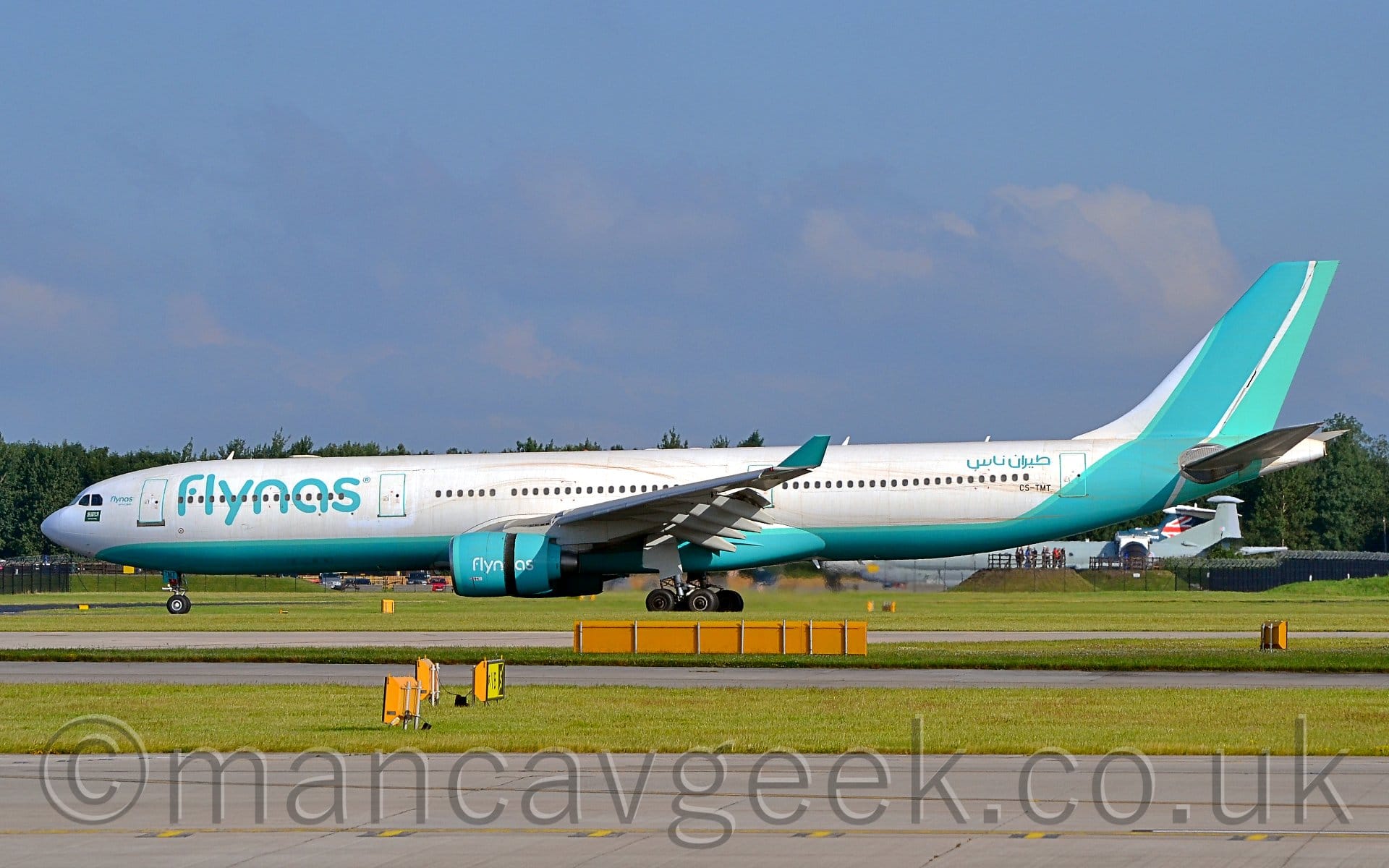 Side view of a twin engined jet airliner slowing down as it moves from right to left, all flaps and spoilers extended from the wings and the thrust reversers activated on the rear of the engine pods, suggesting it has just landed. The plane is mostly white, with a pale blue belly that turns up to fill the tail. There are large, billboard-style "flynas" titles on the forward fuselage in the same pale blue, with some writing in an Arabic script on the on the upper rear fuselage. The engine pods under the wings are the same pale blue, with white"flynas" titles, the engine cowling split because of the thrust reversers, with the last 2 letters on the rear section. A grey runway and a black taxiway are in the foreground, separated by a wide strip of grass. Trees can be seen in the background on the left of the frame, with blue sky with fluffs of cloud filling the rest.