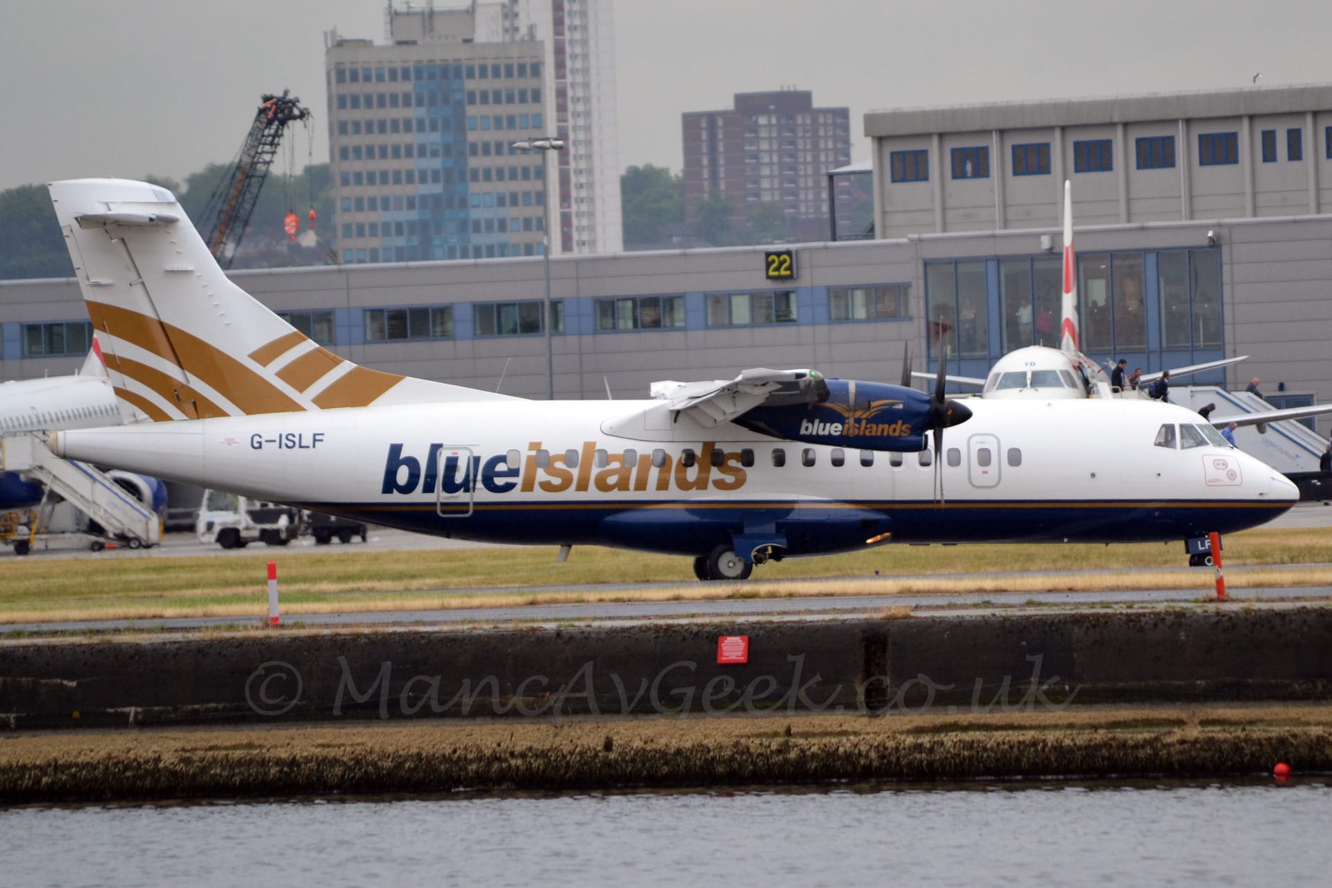 Side view of a high-winged, twin propellor-engined airliner taxiing from left to right along a grey taxiway. The plane is mostly white, with a dark blue belly and a golden pinstripe. There are blue and gold "Blue Islands" titles on the lower rear fuselage. The engine pods on the wings are dark blue, with white and gold "Blue Islands" titles under a stylised golden pair of wings extended in flight. A larger version of the middle of these golden wings appears on the white tail. In the foreground, the runway is elevated several metres above a large body of water, while a grey terminal building stretches across the background, with a couple of white jet airliners parked facing the camera in front. A pair of tower blocks stretch into the grey sky beyond.