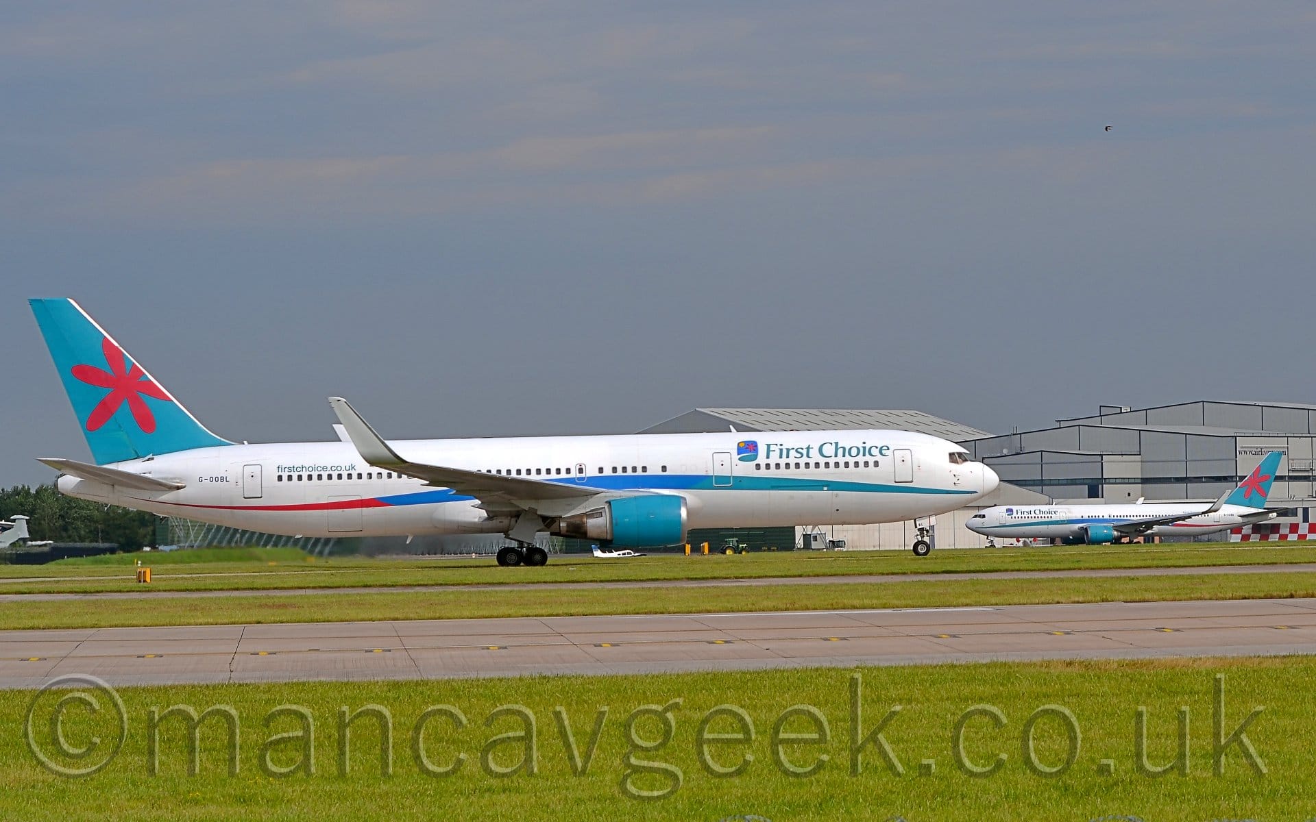 Side view of a twin engined jet airliner moving from left to right as it accelerates along a runway. The plane is mostly white, with blue-green "First Choice" titles on the upper forward fuselage, next to  squar-ish logo that looks like a childs drawing of a holiday destination - blue sky and green ground separated by a white curve, with a yellow sun in the top left corner, and a red asterisk that I think is supposed to be some kind of flower in the top right. A multi-coloured stripe runs along the body, blue-green at the front, blue in the middle, and red at the rear, swooping up slightly over the wing. The tail is a different shade of blue-green, with a red asterisk/flower over the top. The engine pods under the wings are also a slightly different shade of blue green, while they appear to have missed a trick on the up-turned wing-tips, which are just white. Lush green grass fills the foreground, with a grey runway and a pair of taxiways running through. A pair of large grey hangars dominate the background on the right of the frame, with a plane very similar to this one taxiing in front of them in the opposite direction. Blue sky with wisps of grey cloud fill the rest of the frame.