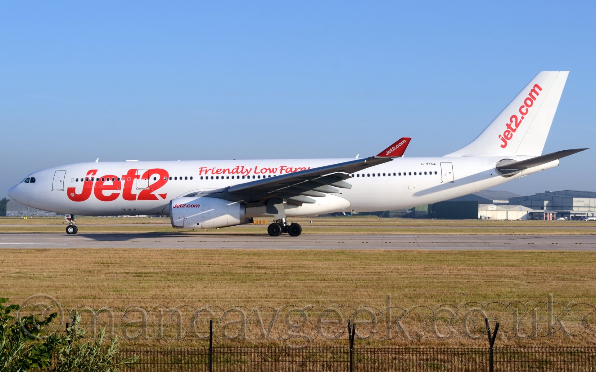 Side view of a twin engined jet aircraft moving slowly from right to left as it starts it's take-off roll. The plane is almost entirely white, with billboard-style red "Jet2" titles on the front fuselage, smaller "Friendly Low Fares" additional text on the upper fuselage over the wings, and red "Jet2.com"  titles diagonally on the tail, and in a small form at the front of the engine cowling under the wings. The up-turned wing-tips are red, with white "Jet2.com" titles. Hot, dry weather has left the green grass lining the sides of the runway in the foreground looking more yellow, even brown in places, with a black metal fence topped with rolls of razor-wire at the bottom of the frame. More grass in the background leads up to large grey hangars in the background, under the plane's tail, with blue sky filling the rest of the frame.