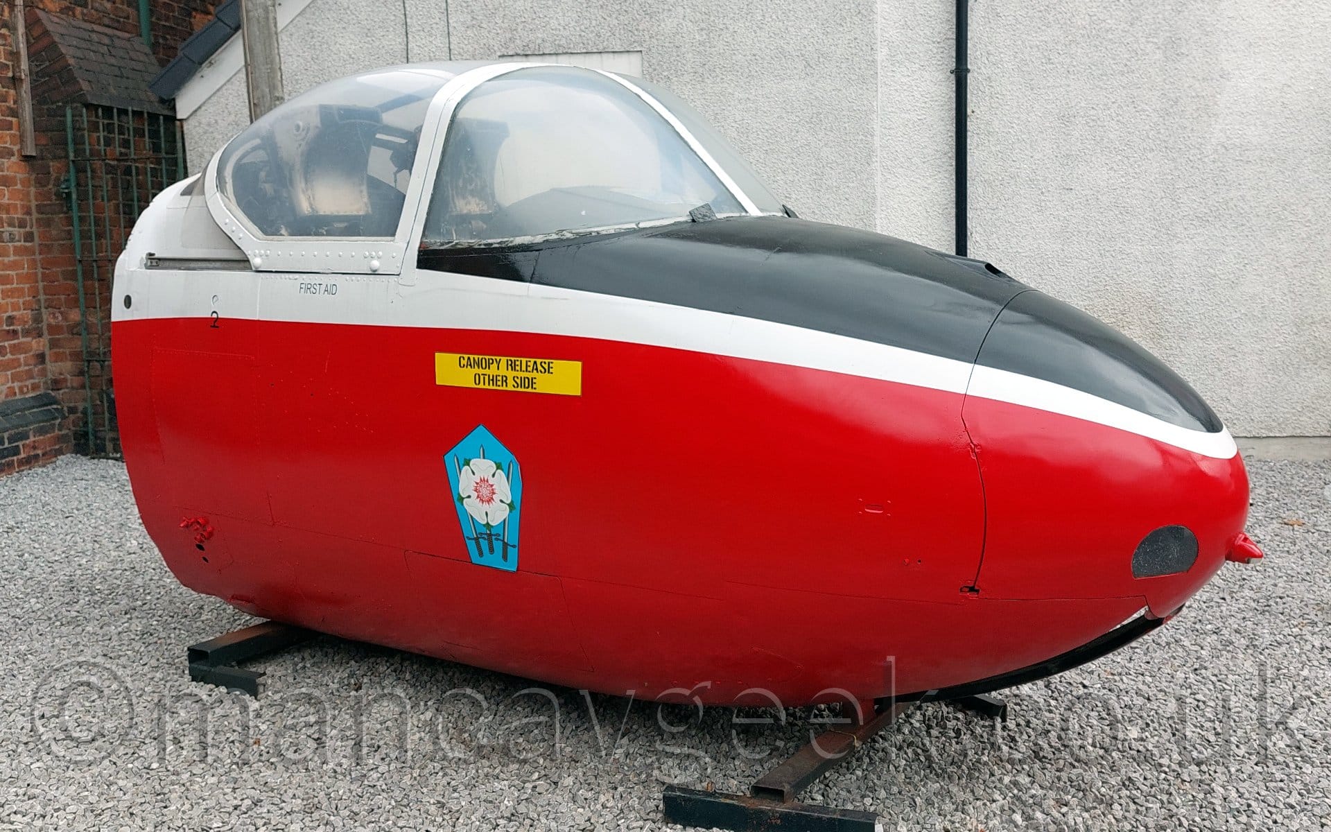 Side view of the forward fuselage and cockpit of a single engined military jet trainer aircraft, sitting on a couple of metal supports on a white gravel base, facing to the right. The plane is mostly red, with a white upper fuselage and frame around the cockpit, and a black anti-glare panel on top of the nose, directly in front of the cockpit. On the side of the nose is a blue pentagon with a white rose in the middle, below a yellow rectangle with the words "Canopy release other side" in black. The white wall of a building fills the background.
