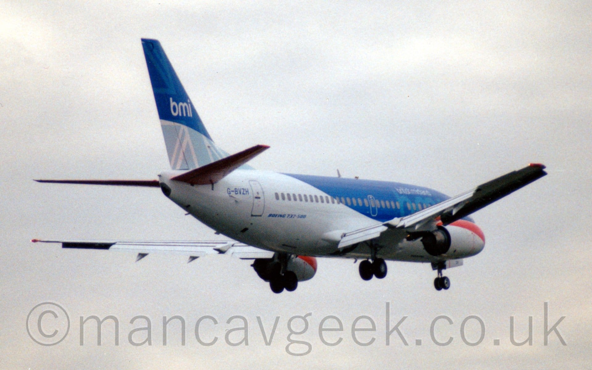 Rear side view of a twin engined jet airliner flying from left to right at a very low altitude, with undercarriage lowered and flaps extended from behind the wings, suggesting it is about to land. The plane is mostly white, with blue forward fuselage with light blue "BMI" and white "British Midland" titles on the forward fuselage. The tail has a modified Union Flag in various shades of blue, with white "BMI" titles. The engines under the wings are white, with a red band around the upper leading edge. Flat white sky fills the rest of the frame.
