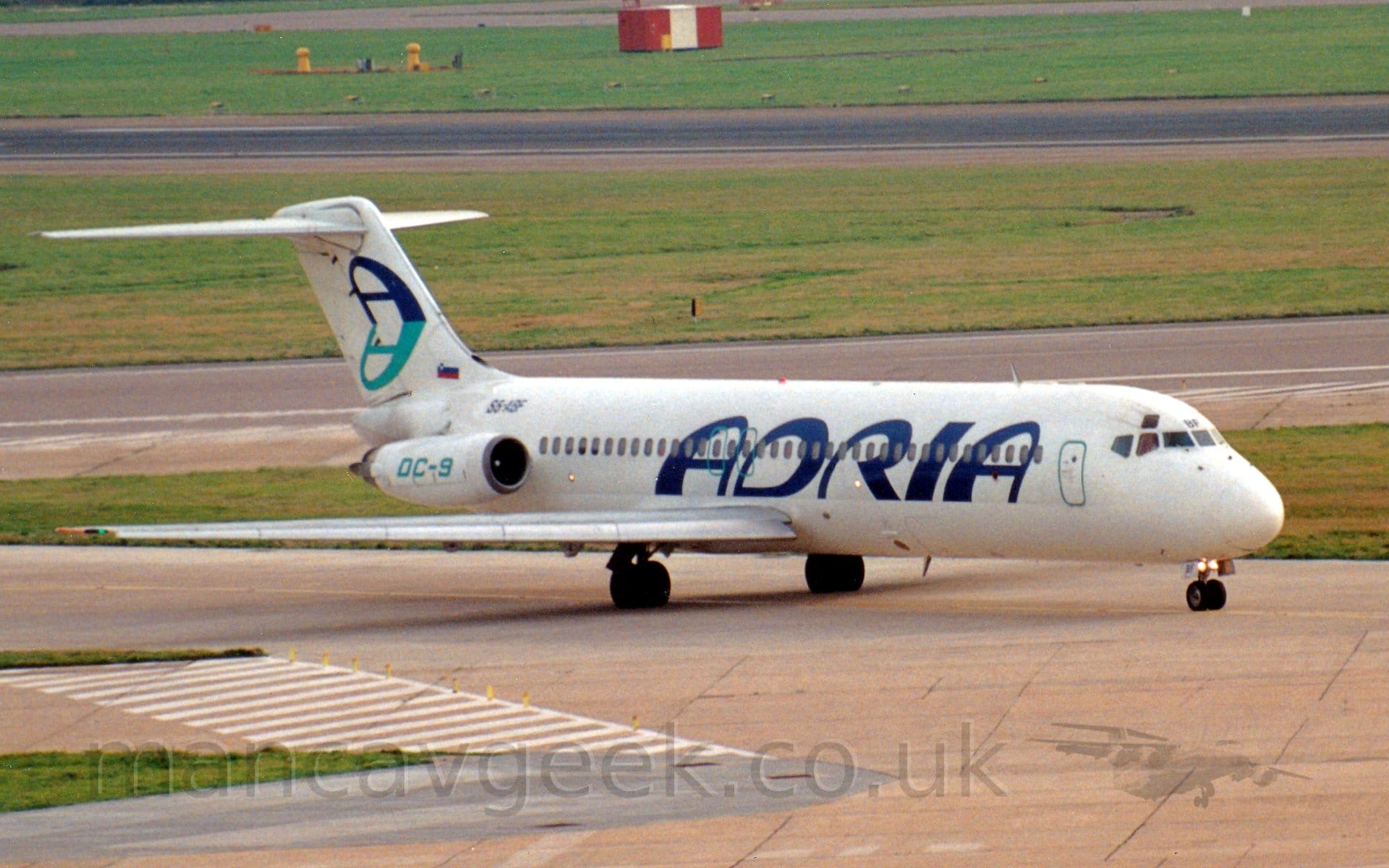 Side view of a twin engined jet airliner with the engines mounted on the sides of the rear fuselage. The plane is mostly white, with dark blue "Adria" titles on the side of the forward fuselage. There is blue-green "DC-9" text on the sides of the engine pods, with the black registration "S5-ABF" on the side of the fuselage just above the engines. The white tail has a large, dark blue upper-case letter "A", with a blue-green reflection at it's base. A pair of bright lights are attached to the top of the nose-wheel leg. Grey concrete apron fills the foreground, with large grassed areas separated by taxiways and a runway in the background.