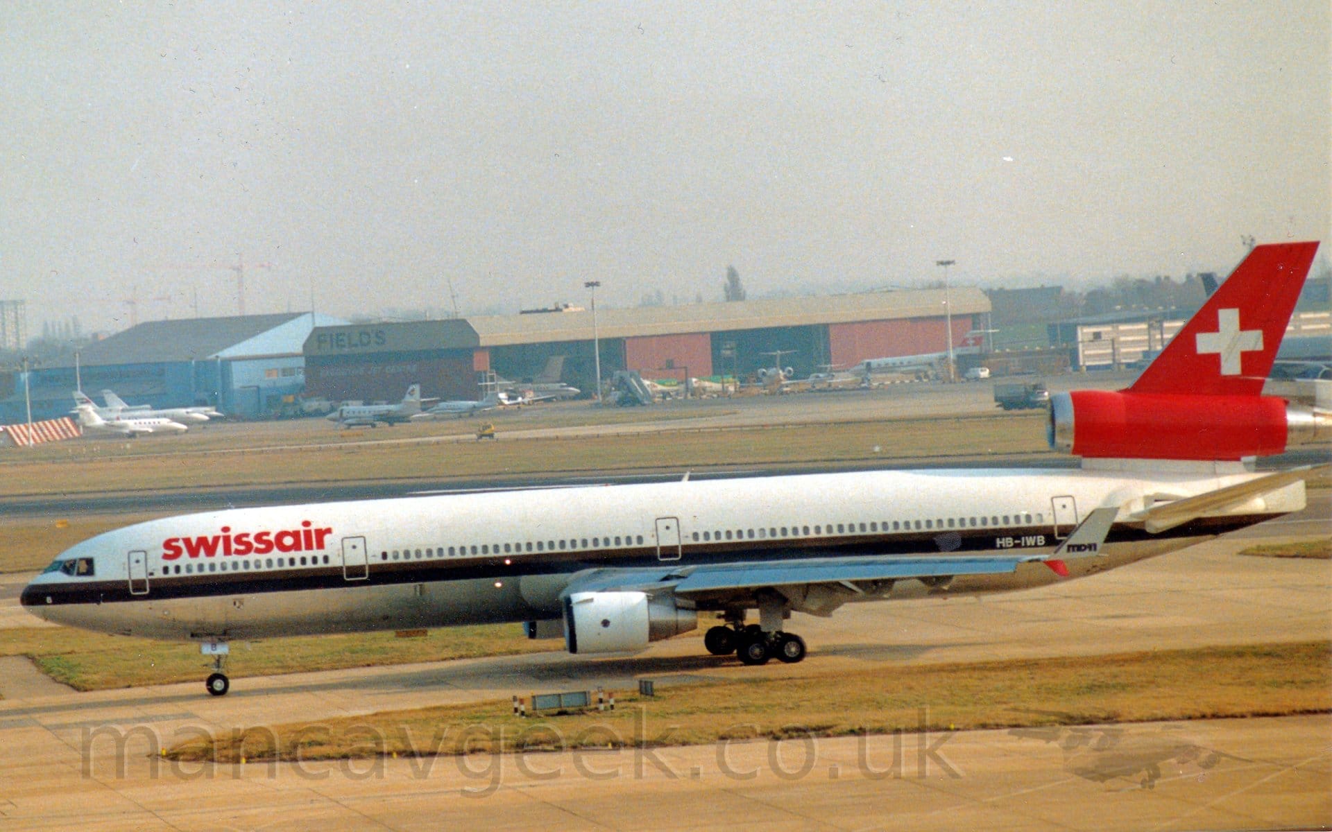 Side view of a 3 engined jet airliner taxiing from right to left. The plane has a white upper fuselage, with a grey belly, a brown and black stripe running the length of the body separating the 2 colours. There are red "SwissAir" titles on the upper forward fuselage, and the white registration "HB-IWB" on the black part of the stripe on the rear fuselage. The tail and centre engine are red, with a large white cross in the middle of the tail. The other engines, mounted under the wings, are white. The white up-turned wingtips have black "MD11" text. Grey concrete taxiways and yellowing grass fill the foreground, with more grass leading up  to a pair of large hangars in the distance, one red, the other blue, with several BizJets parked outside. Hazy grey sky fills the rest of the frame.