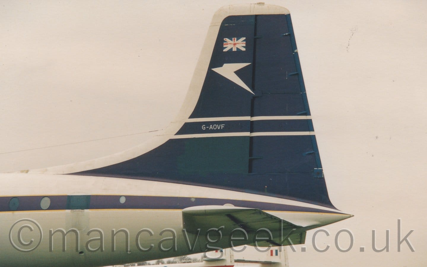 Close-up of the tail of a 1950's-era airliner parked facing to the left. The plane is mostly white, with a thick grey stripe running along the body, covering the passenger cabin windows. The tail is the same dark blue, the leading edge outlined in white, with a pair of thin white stripes along the middle, with the white registration "G-AOVF" in between them. There is a heavily stylised, angular, white flying bird on the upper left of the tail, under a rather faded Union Flag. Flat white sky fills the rest of the frame.