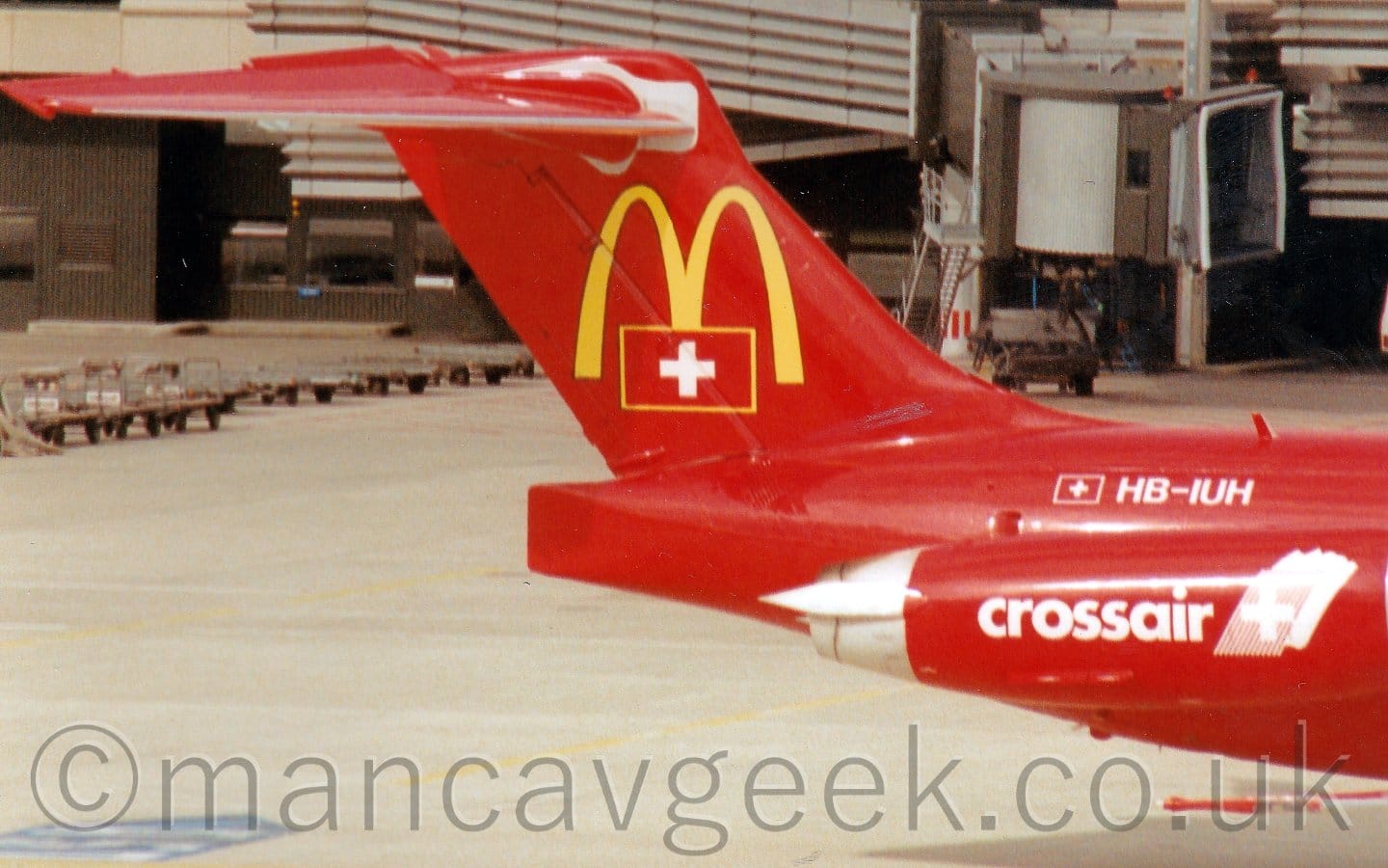 Close-up of the rear fuselage and tail of a twin engined jet airliner with the engines mounted on the sides of the rear fuselage. The plane is mostly red, with a very familiar logo, a pair of yellow arches that resemble a letter "M" on the tail, overlaid at the bottom with Swiss flag (red field with a white cross, outlined in yellow to make it stand out from the rest of the red. A smaller version of this flag, this time outlined in white, is on the upper rear fuselage, next to the white registration "HB-IUH". There are white "CrossAir" titles on the side of the red engine pods below and in front of the tail. Grey concrete apron fills the bottom of the frame, with grey airbridges attached to a grey terminal building in the background at the top.